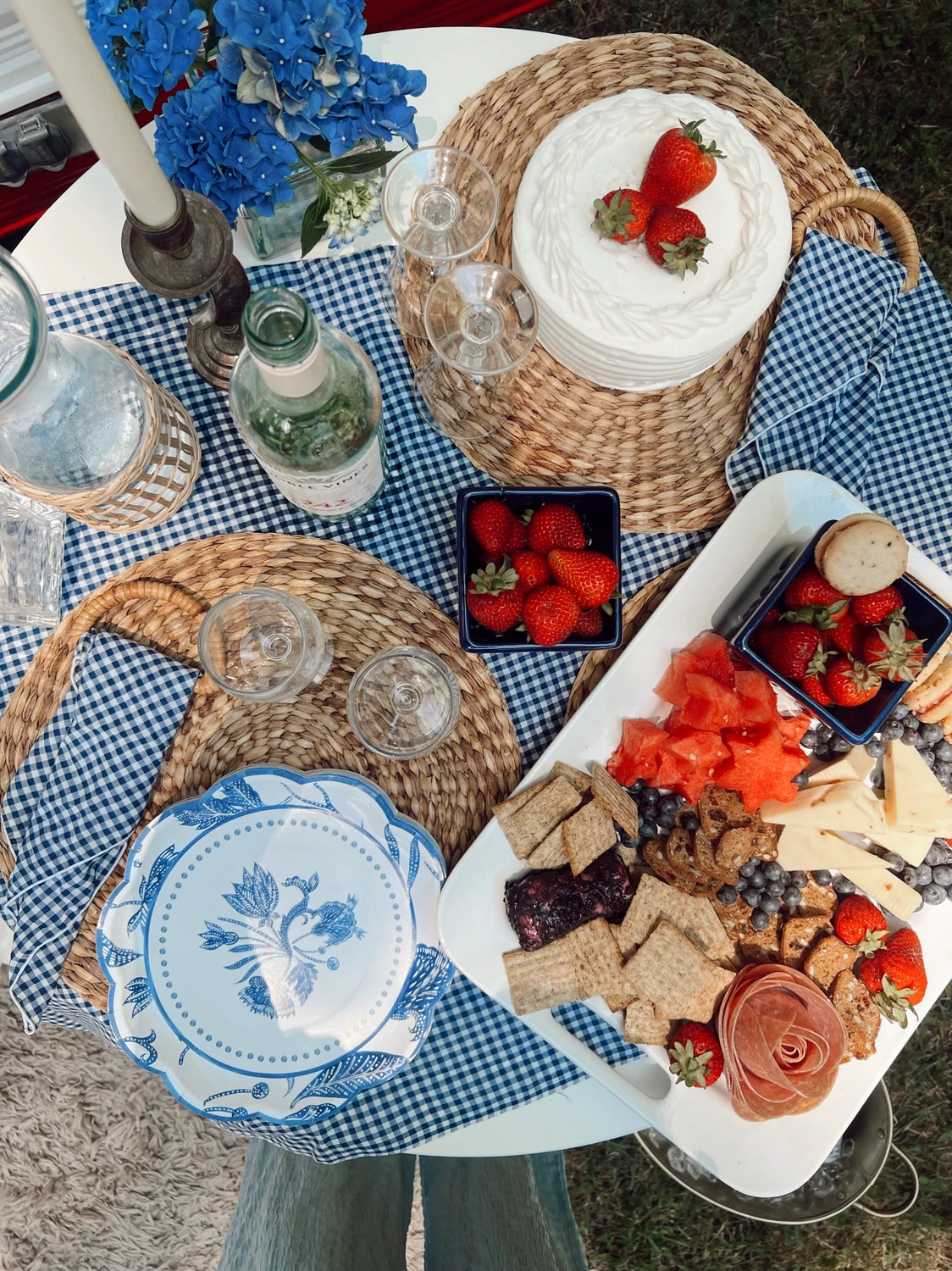 A beautifully arranged overhead shot shows a festive picnic setting with smart 4 july decor using a red, white, and blue color palette. The subtle table decor features a blue-and-white gingham tablecloth, woven rattan placemats, and elegant blue patterned plates. A round white frosted cake topped with fresh strawberries sits near a square blue bowl of strawberries, while a large platter is filled with star-cut watermelon, crackers, cheeses, blueberries, and a meat rose. Completed by fresh blue hydrangeas in a vase, wine glasses, and a bottle of wine, the table setup captures a chic, patriotic summer celebration.