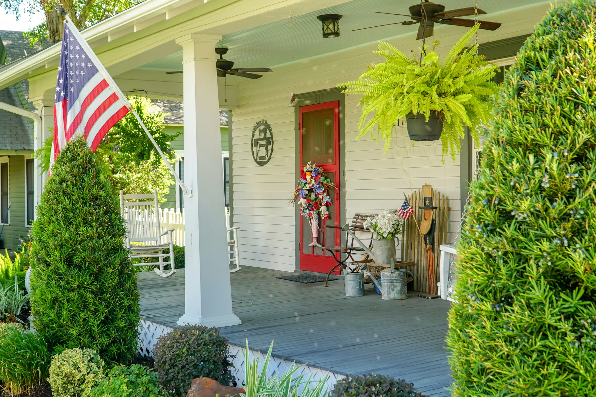 A classic Americana farmhouse porch is decorated with festive front porch decor ideas for summer, celebrating Independence Day with patriotic 4th of july decor. An American flag is proudly displayed from a white pillar, while the bright red screen door features a large, colorful wreath with red, white, and blue accents. A lush, hanging fern adds a pop of vibrant green under the porch ceiling, and vintage metal watering cans and white flowers sit on a rustic bench nearby. The scene is framed by manicured green bushes and white rocking chairs in the background, creating a quintessential and welcoming summer atmosphere.