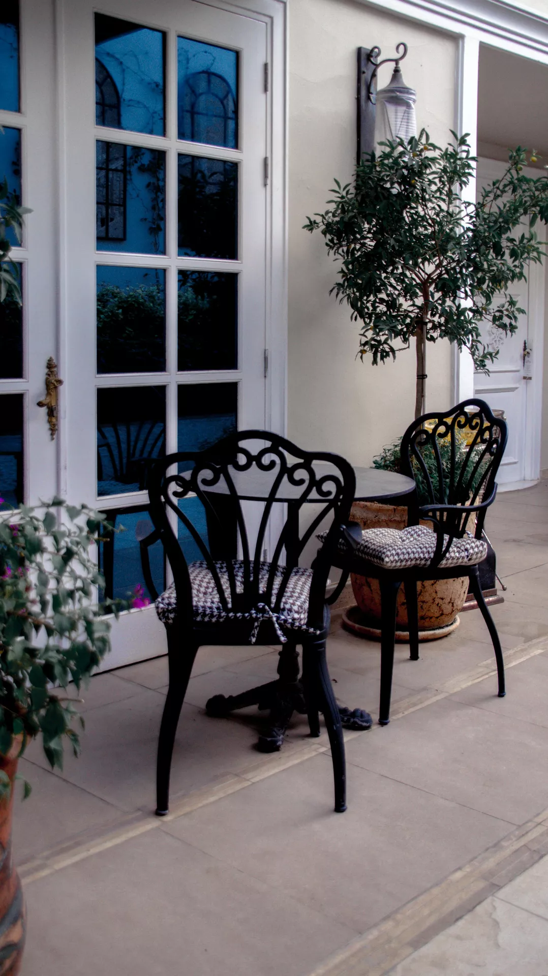 An elegant Mediterranean-style patio features sophisticated front porch decor ideas for summer with a focus on ornate black furniture. A pair of wrought-iron bistro chairs with intricate scrollwork and patterned seat cushions are arranged around a small matching circular table. The seating area is nestled next to a large white French door with glass panes and a tall, slender potted tree in a textured terracotta planter. The warm, neutral-toned stone flooring and the soft cream exterior wall create a timeless and inviting outdoor space perfect for a morning coffee or a quiet summer evening.