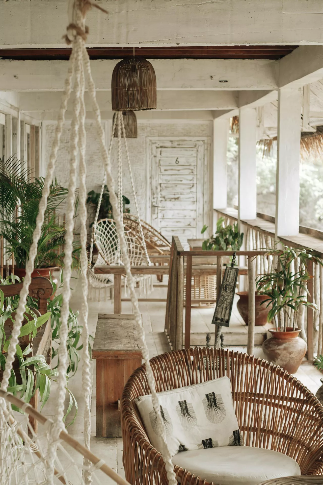 A rustic, weathered wooden veranda showcases tropical front porch decor ideas for summer with strong boho vibes. The space is filled with natural textures, featuring a round rattan armchair with a patterned cushion in the foreground and a white macramé hanging chair suspended by thick woven ropes. Earthy terracotta pots holding lush green palms and tropical plants line the railing, while a dark wicker pendant light hangs from the white-washed ceiling. The distressed white wooden door and aged timber benches enhance the relaxed, bohemian atmosphere of this shaded outdoor retreat.
