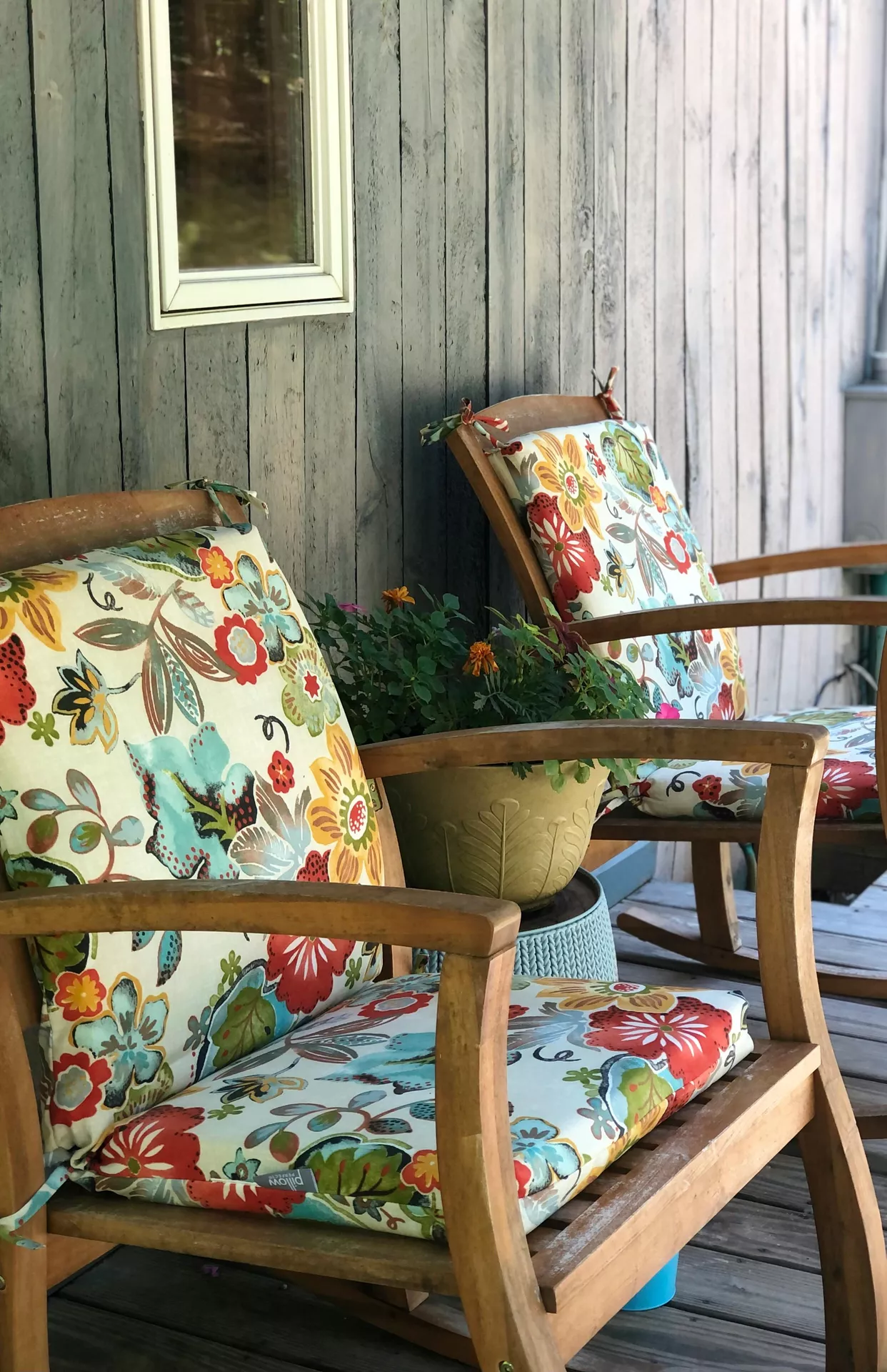 Two wooden rocking chairs sit on a rustic deck, showcasing vibrant front porch decor ideas for summer. Each chair is fitted with thick, colorful seat cushions featuring a bold floral pattern in shades of red, turquoise, orange, and lime green. Between the chairs, a green decorative planter filled with small orange flowers rests on a light blue textured garden stool. The setting is backed by a weathered grey wooden wall, creating a cheerful and inviting outdoor seating area perfect for enjoying a warm sunny day.