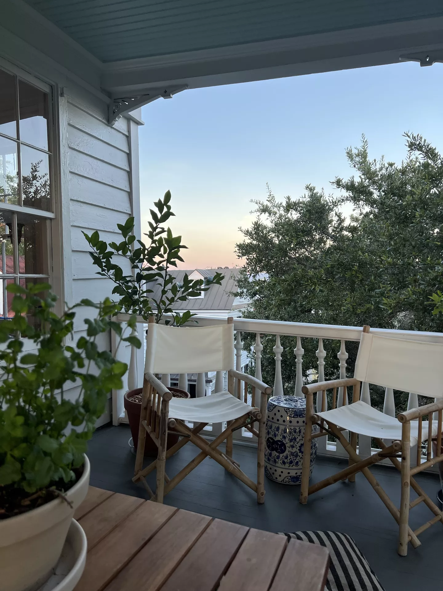 A serene elevated balcony showcases elegant front porch decor ideas for summer, featuring a pair of bamboo director's chairs with white canvas seating. Positioned between the chairs is a classic blue and white ceramic garden stool used as a side table. Lush green potted plants, including a lemon tree in a terracotta pot, add a natural touch to the white-railed porch. The setting is framed by the soft glow of a sunset in the background and a view of neighboring rooftops and large leafy trees, creating a peaceful and sophisticated outdoor retreat.