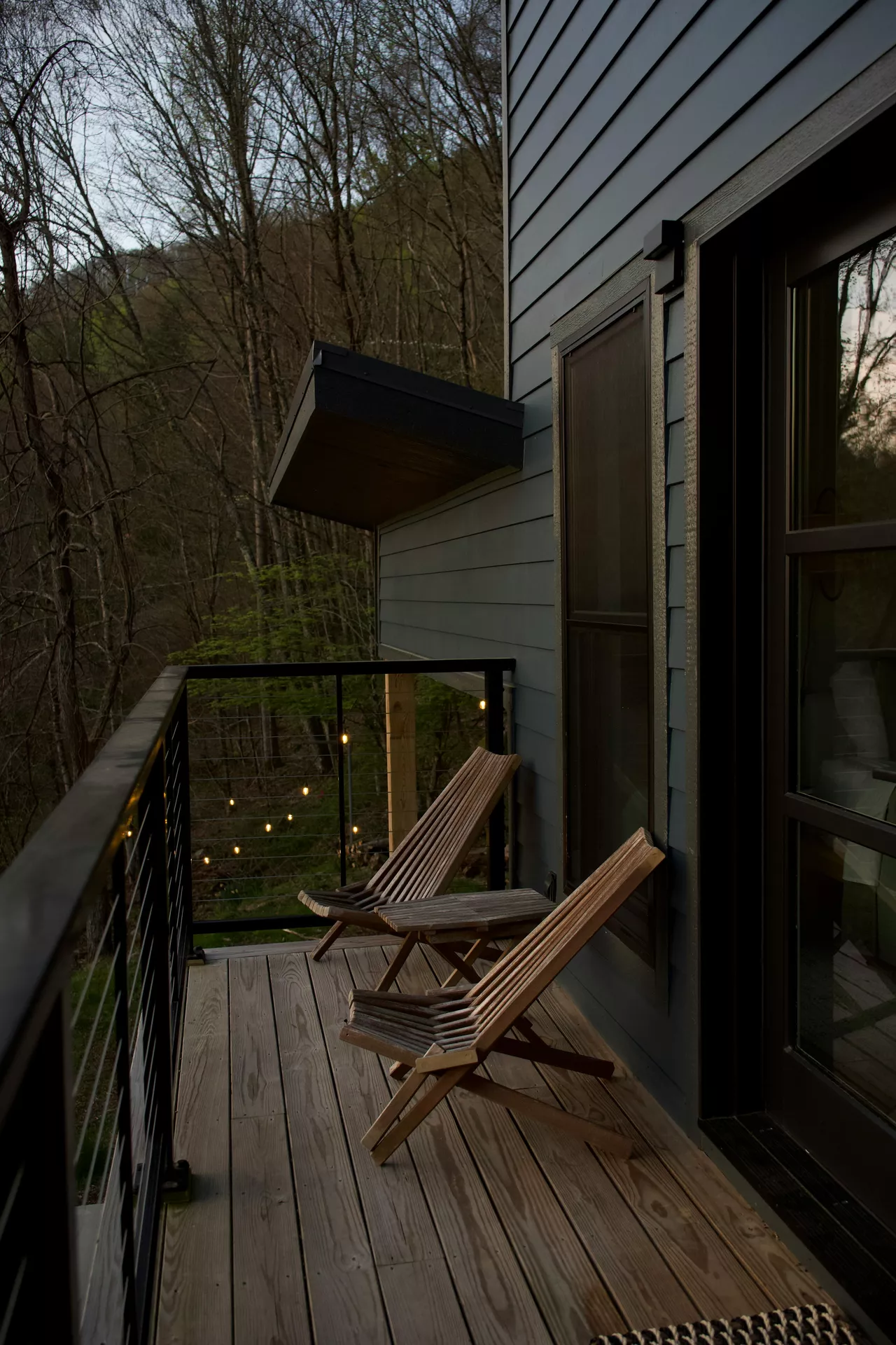 A modern wooden deck overlooks a lush forest at dusk, presenting atmospheric front porch decor ideas for summer. Two slatted wooden lounge chairs face the trees, separated by a small matching side table. In the background, warm fairy lights are strung across the railing and through the branches, creating a magical glow against the darkening woods. The blue siding of the house and the clean lines of the black metal railing contrast with the natural scenery, offering a perfect, tranquil spot for a summer evening outdoors.