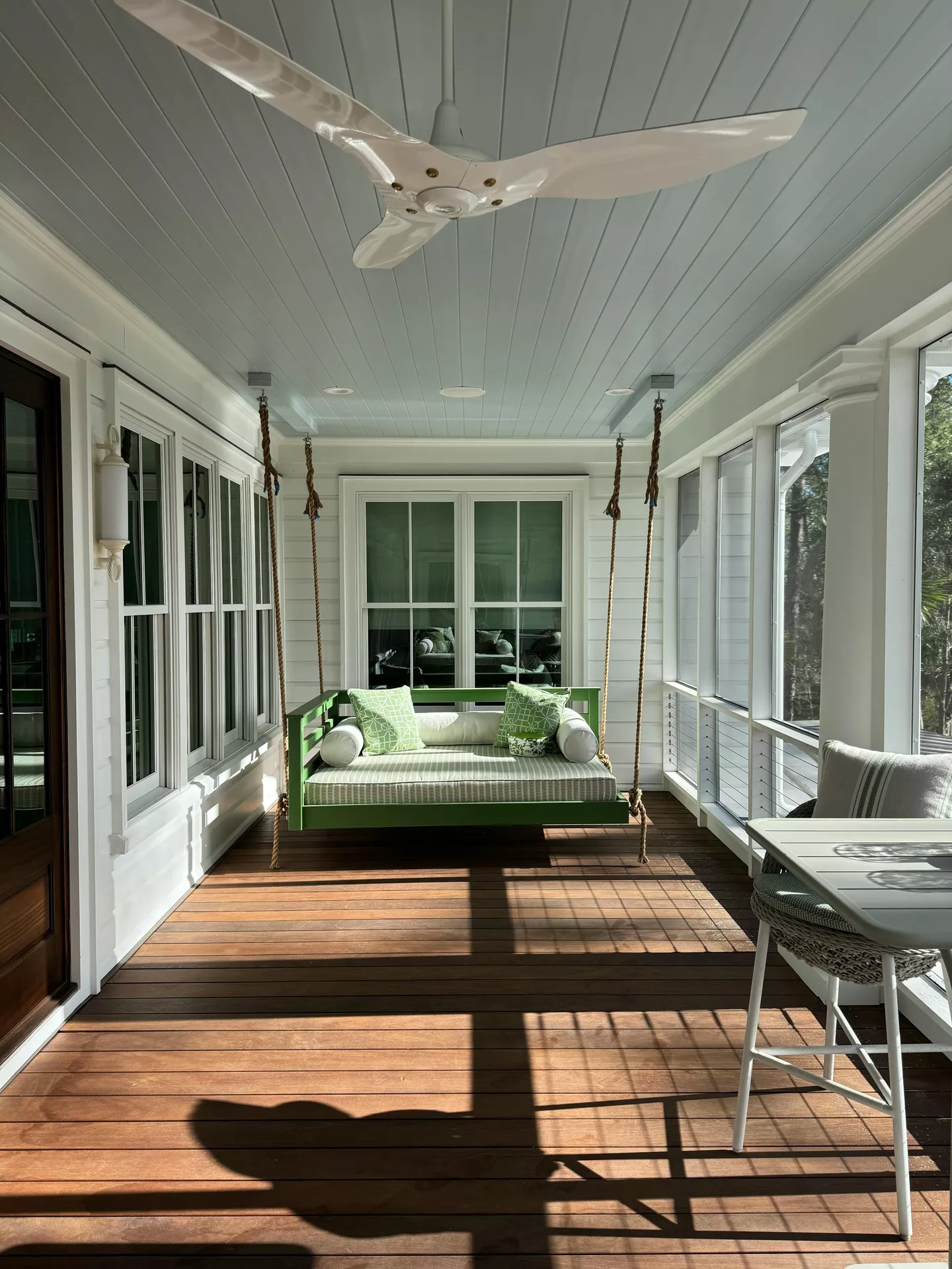 A bright, sunlit screened-in porch displays inviting front porch decor ideas for summer with an emphasis on light and fresh colors. A large, green-painted daybed swing hangs from heavy ropes in the center, adorned with striped and patterned lime green cushions. The space features white shiplap walls, a light blue paneled ceiling with a modern white fan, and warm natural wood flooring. To the right, a white bistro table and woven chair sit near the screened windows, allowing for a clear view of the surrounding greenery while maintaining a cool and airy atmosphere.