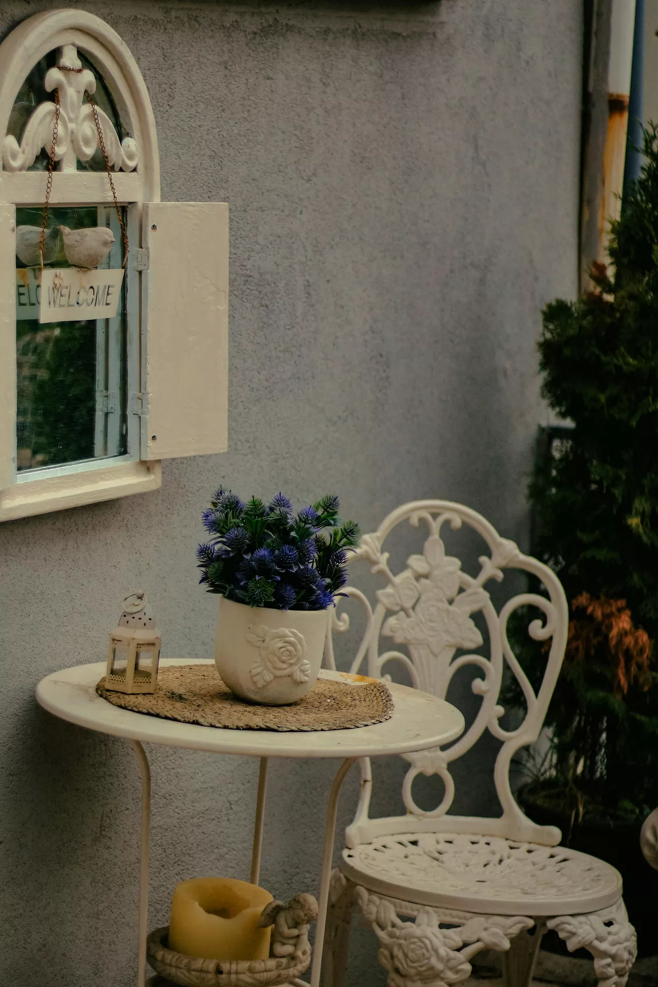 A vintage-style white wrought iron bistro set creates a charming outdoor nook, showcasing inspired front porch decor ideas for summer. The round table is styled with a woven placemat, a small lantern, and a white pot featuring a rose relief filled with deep purple thistles. In the background, a decorative white arched mirror with shutters and a 