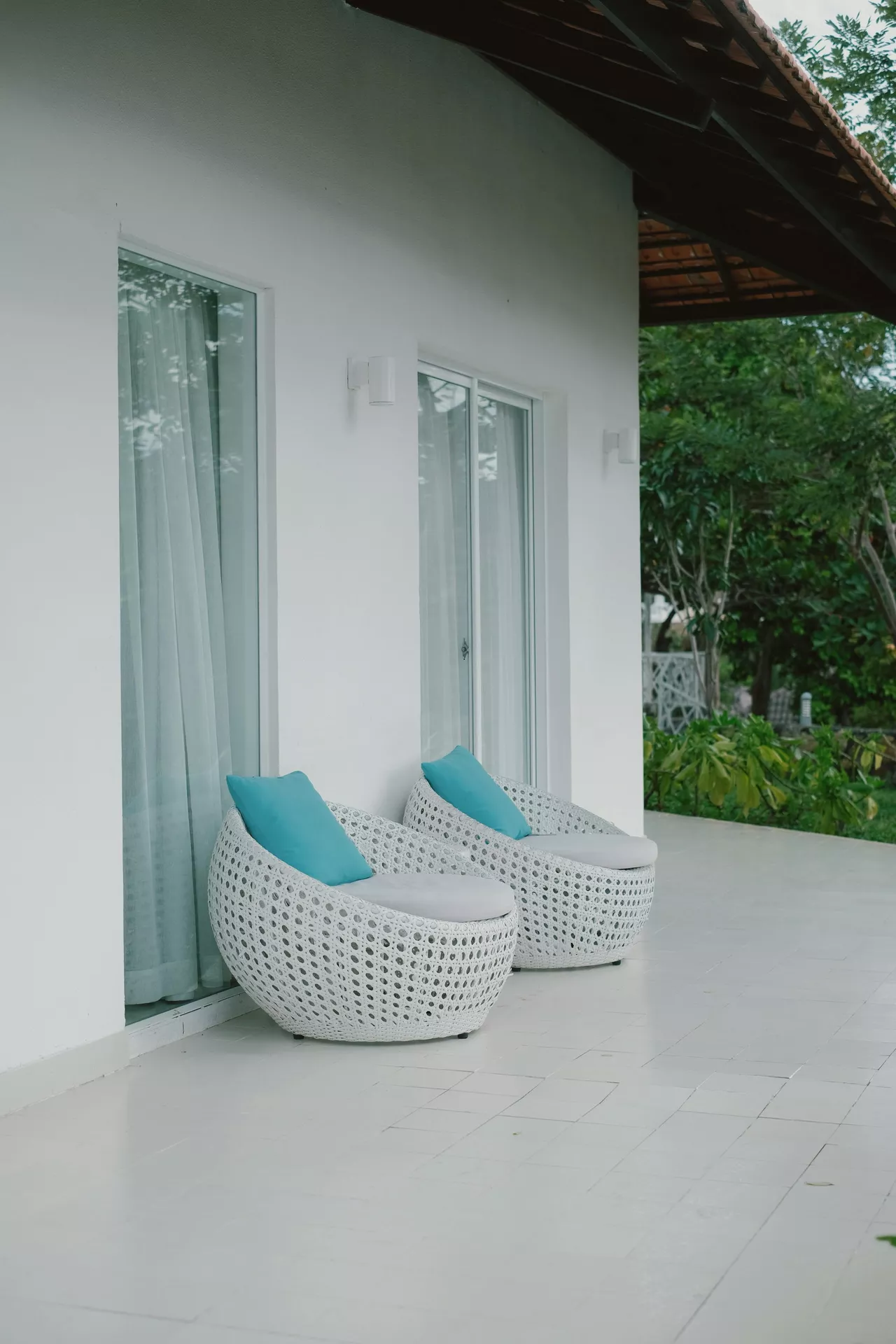 A clean and minimalist white patio features modern front porch decor ideas for summer with two comfortable round beach chairs. These white woven wicker chairs are styled with light grey seat cushions and vibrant turquoise accent pillows, adding a pop of coastal color to the monochrome setting. The chairs are positioned against a bright white exterior wall with large floor-to-ceiling glass doors, creating a fresh and airy outdoor lounge area that perfectly captures a breezy, tropical vacation atmosphere.