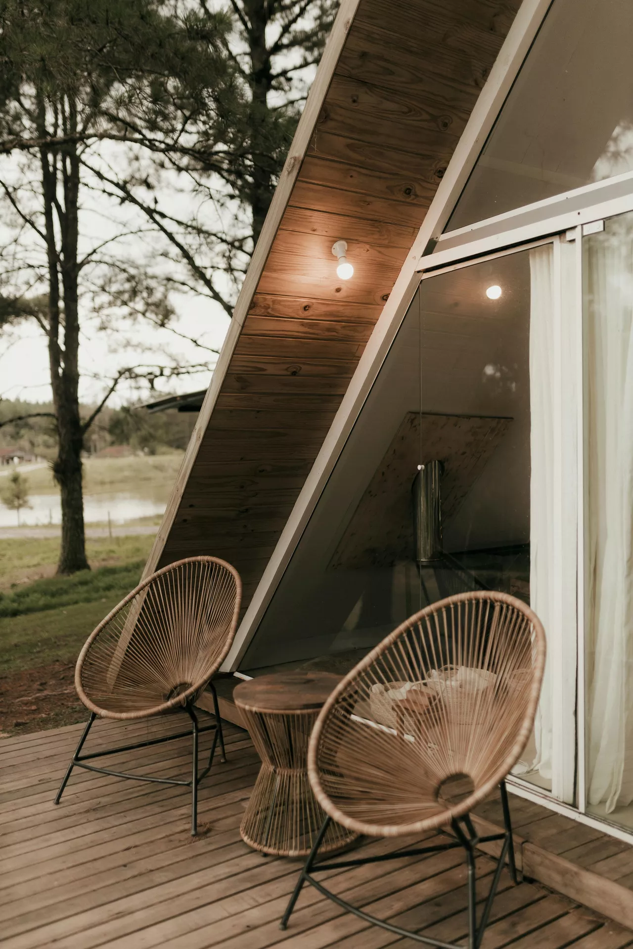 An A-frame wooden cabin features a cozy deck with trendy front porch decor ideas for summer, highlighted by two round string chairs. The tan Acapulco-style chairs are paired with a matching hourglass-shaped side table on the natural wood planks. A warm exterior light bulb glows under the overhanging roof eaves next to large glass sliding doors that reveal a minimalist interior. In the background, tall pine trees and a glimpse of a quiet lake create a peaceful, rustic atmosphere for a summer evening getaway.