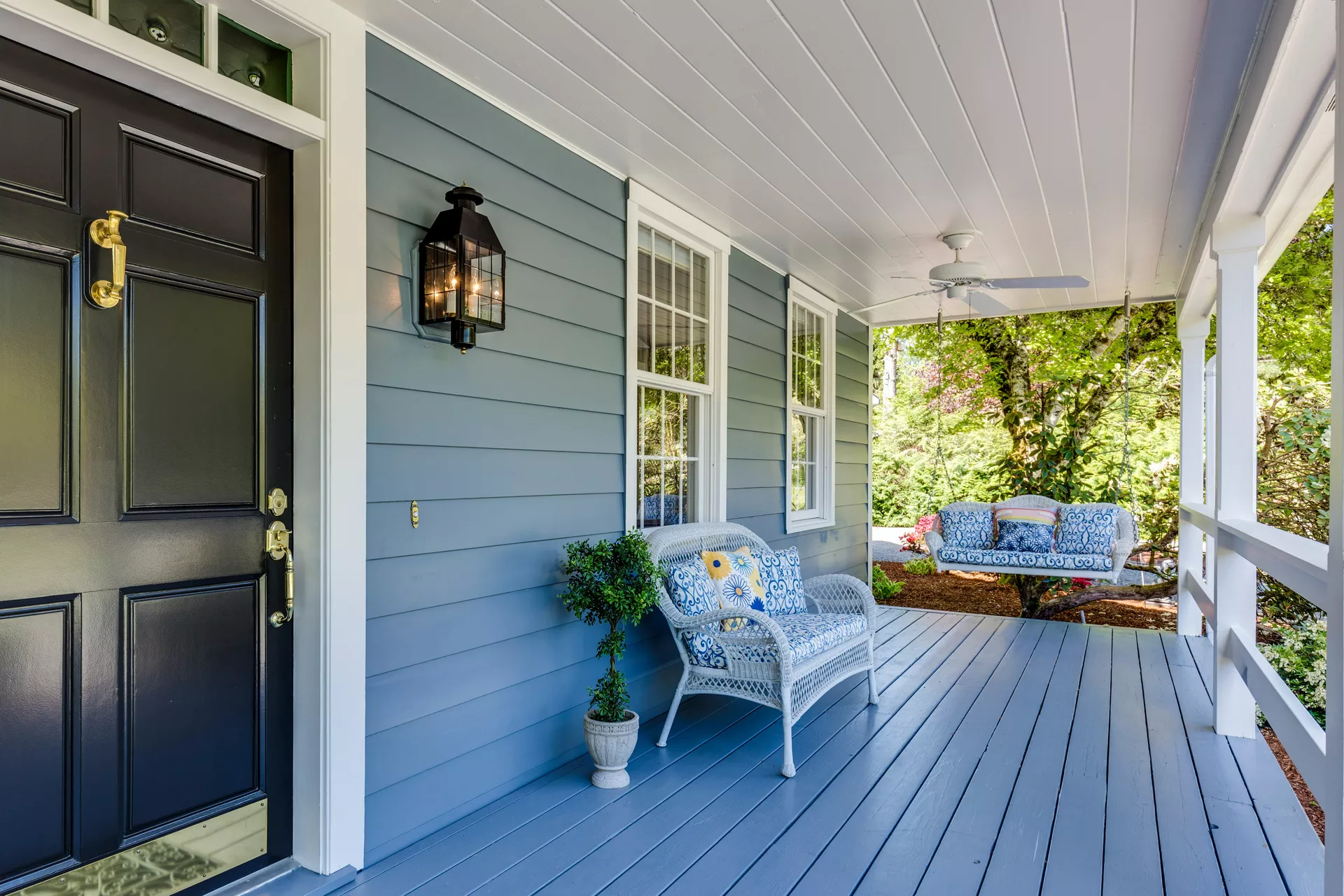 A welcoming porch features elegant front porch decor ideas for summer with a cohesive blue and white color palette. The house is designed with a tone on tone look, matching the blue horizontal siding with the painted wooden deck floor. A black front door with gold hardware stands to the left, while white wicker furniture, including a cushioned armchair and a hanging porch swing, provides a comfortable seating area. Patterned blue and yellow pillows add a splash of color, and a black lantern-style wall light and a white ceiling fan complete the classic, breezy outdoor living space.