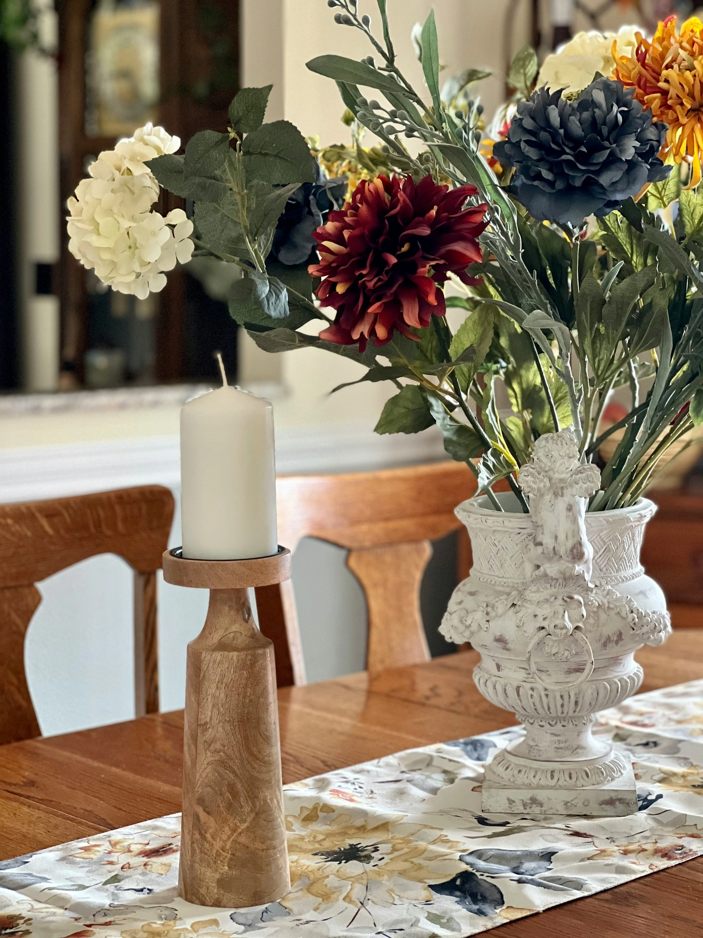 An elegant dining room setup features rich kitchen table decor ideas for summer arranged along a floral-patterned fabric table runner. A stately, white distressed vintage urn serves as a classical planter, holding a dense silk flower arrangement with prominent burgundy and navy dahlia's, cream hydrangeas, yellow chrysanthemums, and leafy green stems. Standing alongside the ornate vase on the wooden table is a tall, carved wooden pillar candle holder supporting a thick white unlit candle. In the background, matching wooden dining chairs complete the warm, sophisticated, and inviting summer table display.