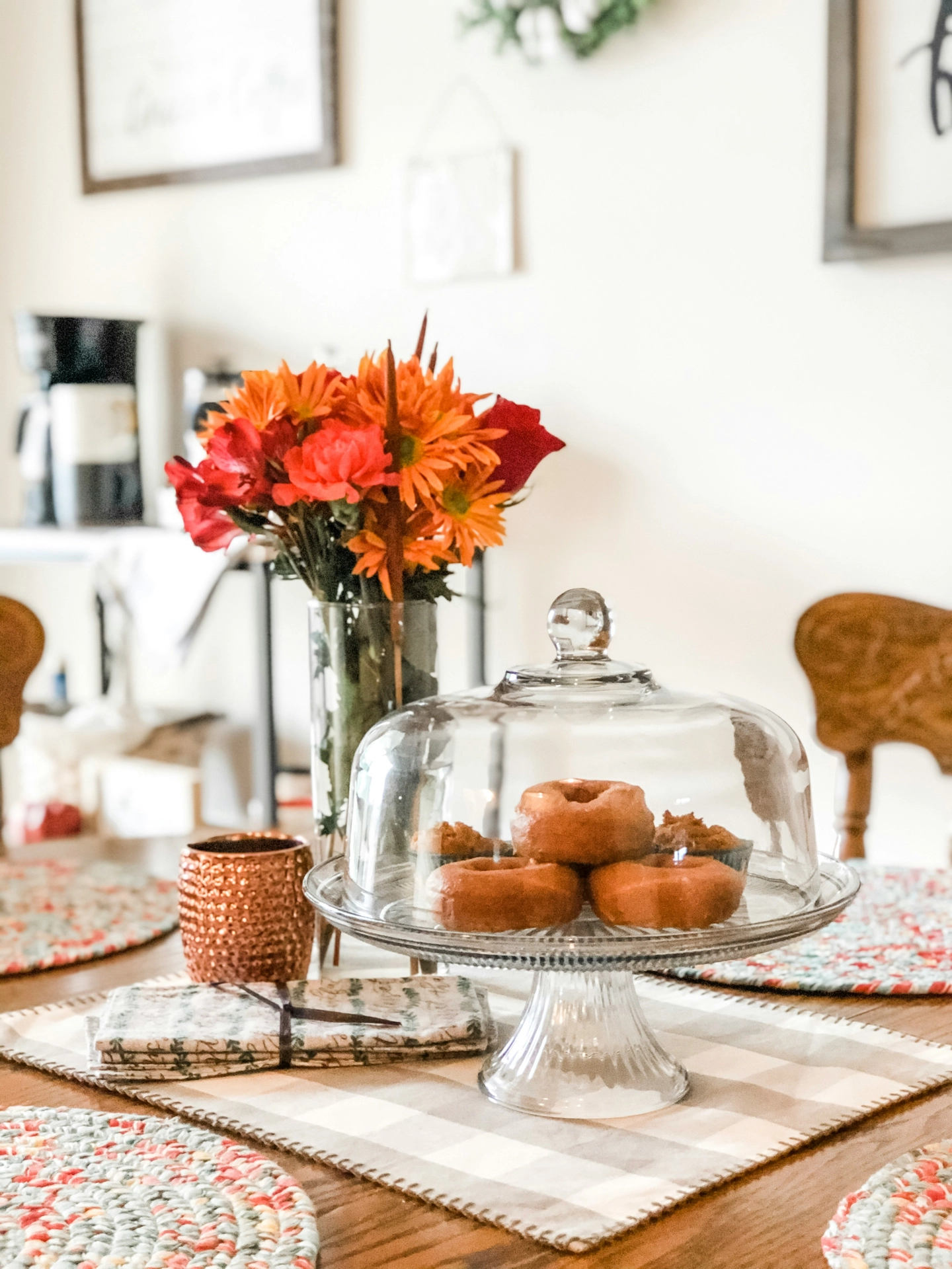 A warm, inviting breakfast setting showcases delightful kitchen table decor ideas for summer atop a wooden dining table. In the center, a clear glass cake stand with a dome lid keeps a tempting stack of chrysantemums, alstroemeria and pastries fresh, featuring golden-brown glazed donuts. Directly behind the treats, a tall glass vase holds a vibrant floral bouquet filled with bright orange chrysanthemums and deep red alstroemeria lilies. The table is beautifully arranged with a beige checkered runner, a textured copper cup, folded napkins, and colorful braided round placemats, creating a cozy and cheerful summer morning atmosphere.
