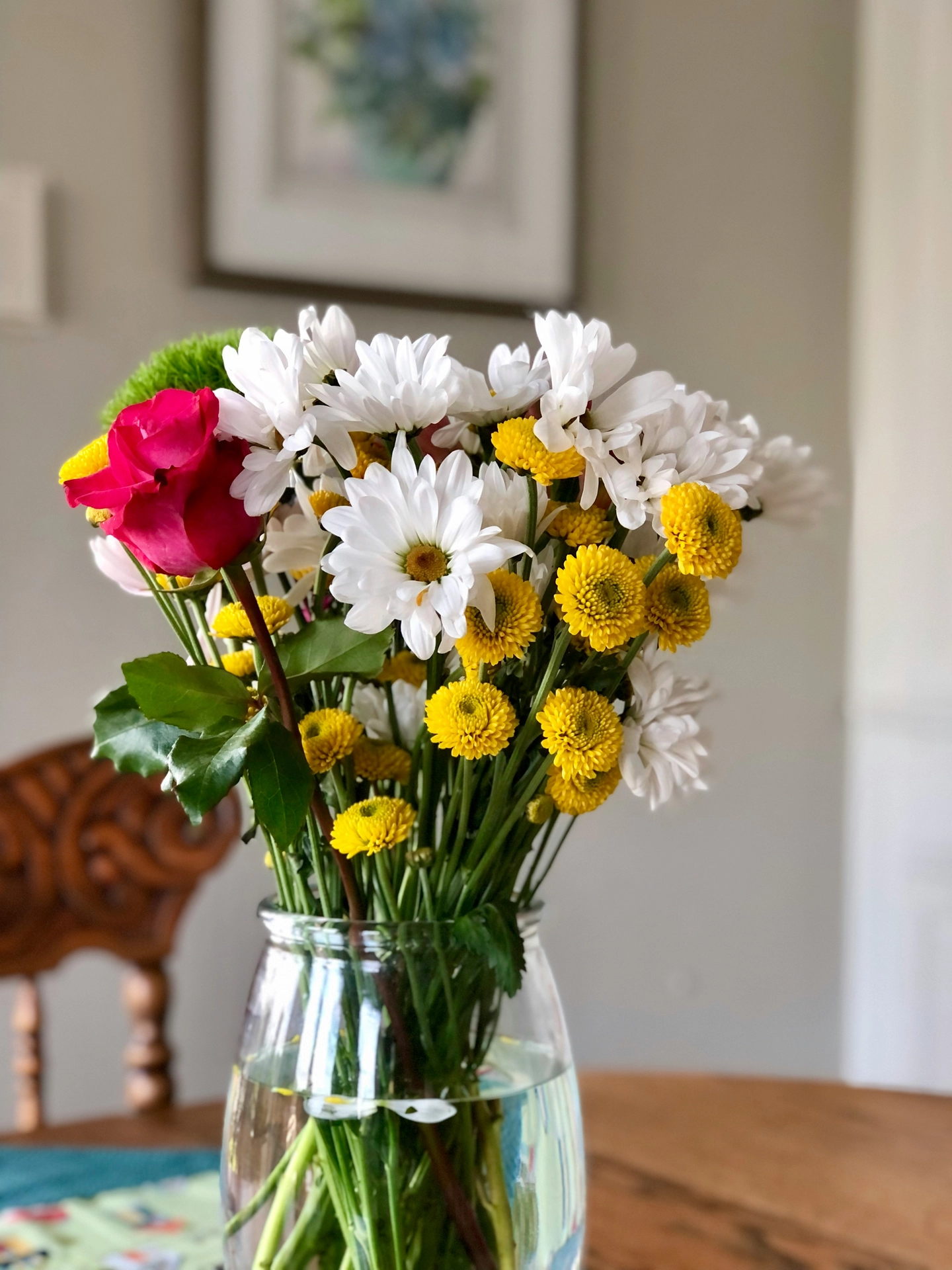 A bright close-up shot presents lively kitchen table decor ideas for summer with a fresh, cheerful floral display. A clear glass vase filled with water holds a colorful mixed bouquet of daisies and crysanthemums, showcasing crisp white daisy petals with yellow centers alongside small, round yellow button chrysanthemums. A single vibrant hot pink rose adds a bold splash of color to the left side of the arrangement, which is surrounded by fresh green leaves and stems. In the soft-focus background, a wooden dining chair and a framed picture on a neutral wall complete the inviting, cozy indoor summer setting.
