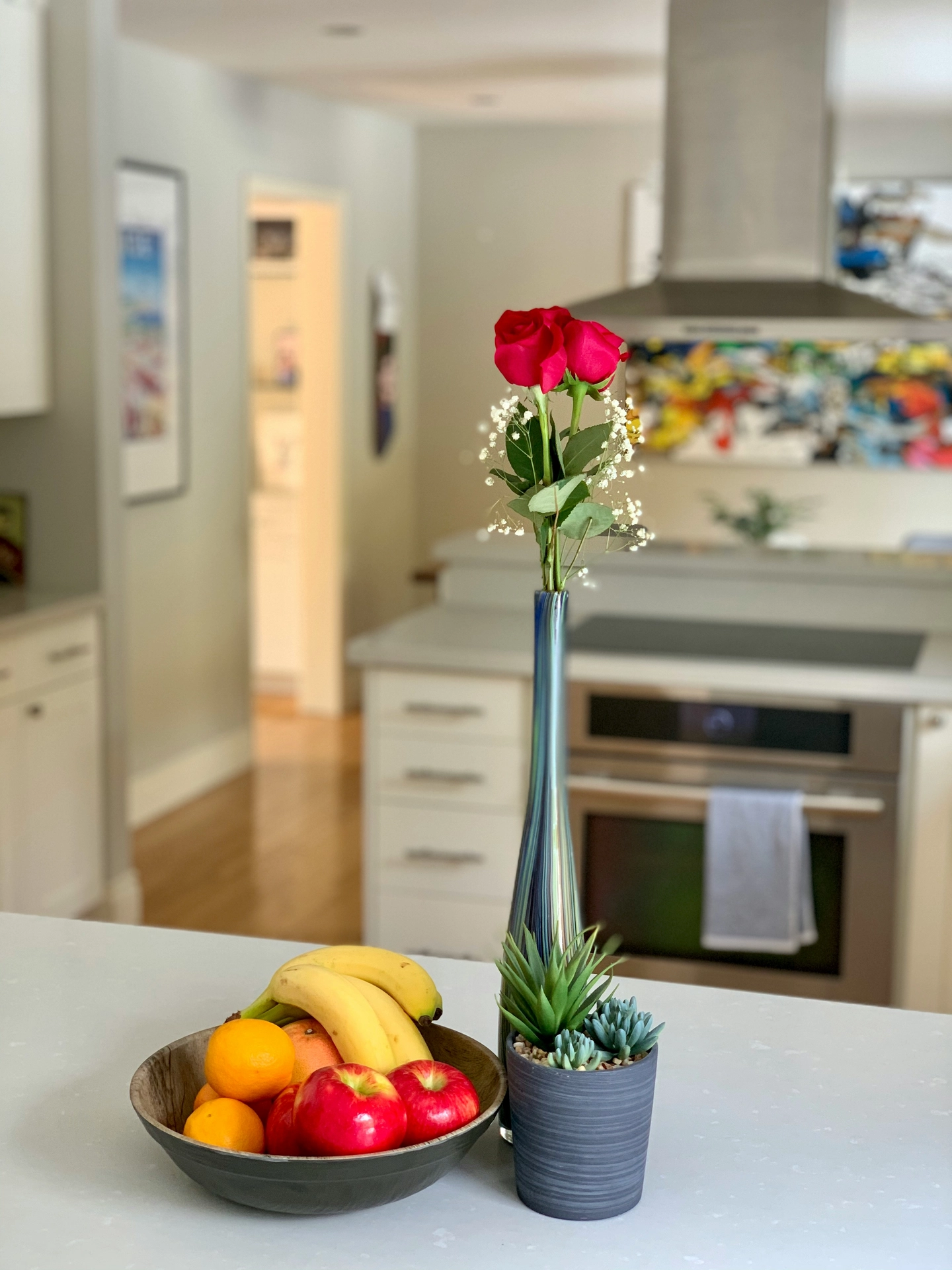A modern and bright countertop arrangement showcases simple kitchen table decor ideas for summer with natural, vibrant accents. The center setup features a beautiful fruit bowl and red rose pairing, with a dark wooden bowl filled with yellow bananas, bright red apples, and oranges. Next to the fruit, a very tall, slender blue-and-green iridescent glass bud vase holds fresh red roses accented with delicate white baby's breath, while a small dark grey pot with faux green succulents sits at the base. In the blurred background, a contemporary kitchen with white cabinets and stainless steel appliances enhances the clean, summery feel.