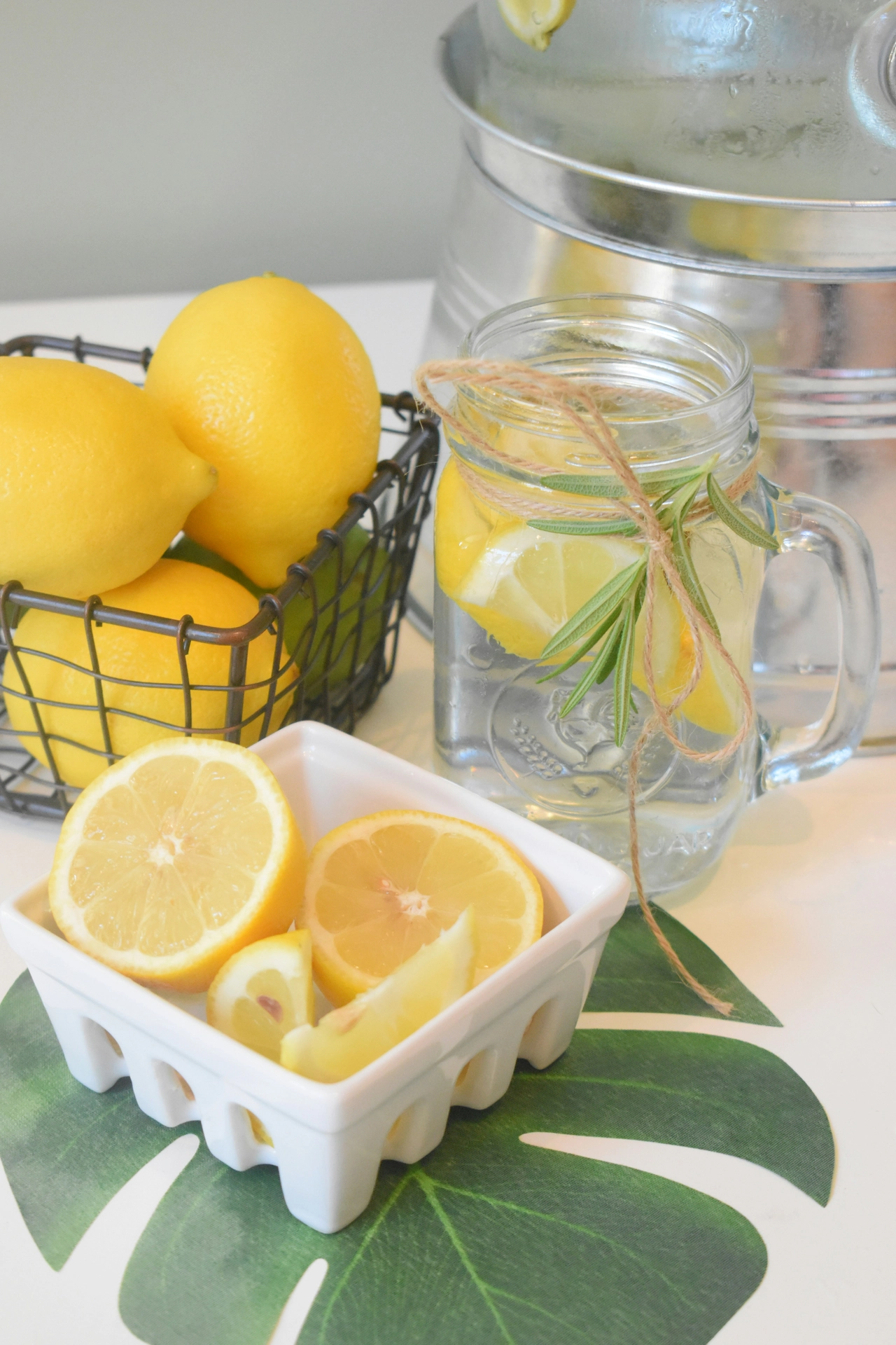 A vibrant close-up setup showcases refreshing kitchen table decor ideas for summer using bright citrus accents. A clear glass mason jar mug is filled with iced water, lemon slices, and a sprig of rosemary, tied neatly with a simple twine bow. Next to the drink, a white ceramic berry basket holds freshly cut lemon halves and wedges, resting on a green monstera leaf placemat. In the background, a dark wire basket is filled with whole lemons and limes, sitting next to a large stainless steel beverage chiller, creating a crisp, thirst-quenching, and sunny summer display.