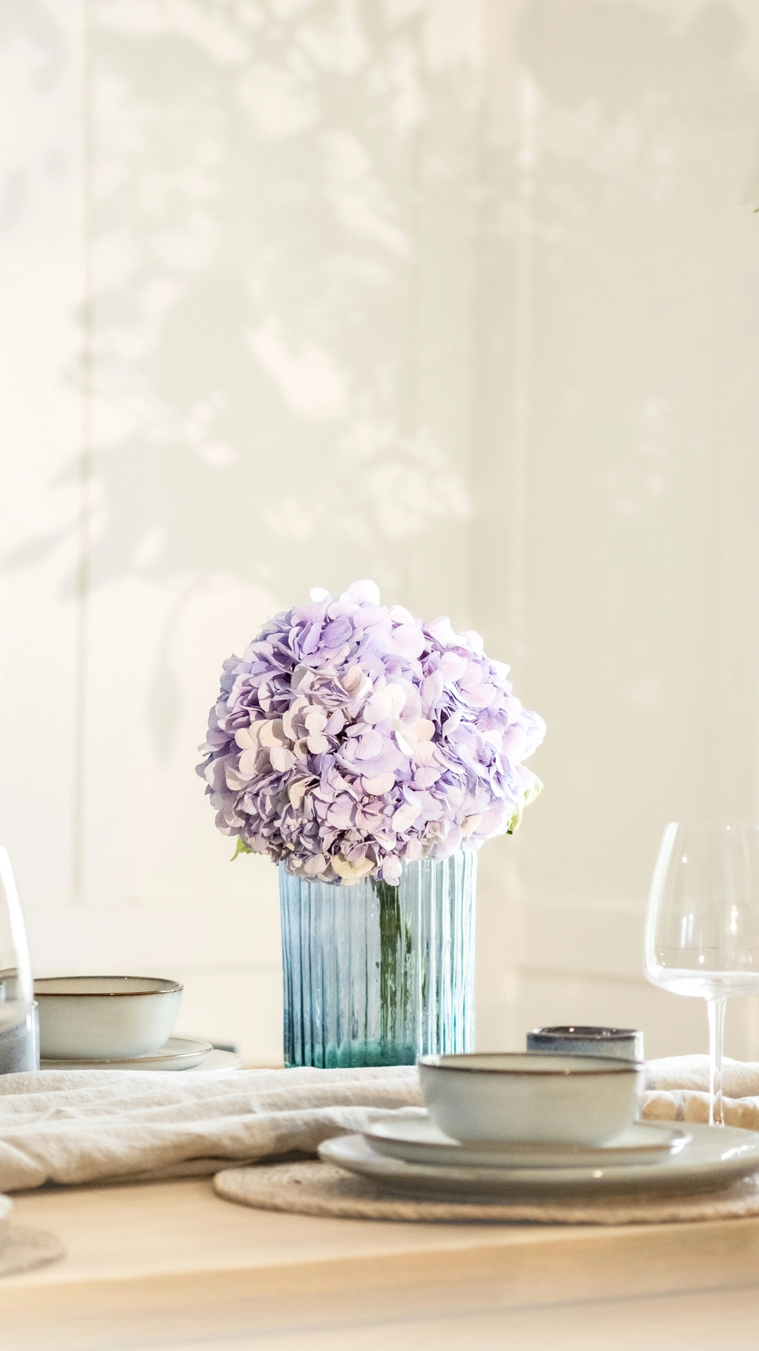 A bright and elegant dining setting showcases organic kitchen table decor ideas for summer with a dreamy color palette. A ribbed, light blue translucent glass vase stands as the centerpiece, holding a large, full bloom of a light purple hydrangea with delicate violet and cream petals. The wooden table is beautifully arranged for a meal with woven round placemats, minimalist light grey ceramic dinnerware, wine glasses, and a softly textured neutral table runner. Artistic leafy shadows are cast by warm sunlight across the clean white paneled wall in the background, creating a serene and breezy summer atmosphere.