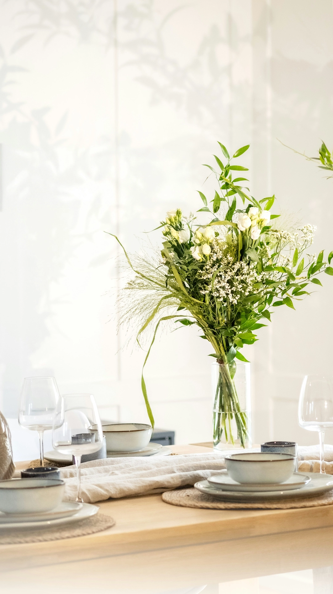 A sunlit dining room features elegant kitchen table decor ideas for summer with a natural, airy table setting. Standing in a clear cylinder glass vase is a textured lisianthus & baby's breath bouquet mixed with long green leafy stems and delicate ornamental grasses. The light wood dining table is set for a meal, featuring round woven placemats, light grey ceramic plates and bowls, wine glasses, and a crumpled beige linen table runner. Soft, dappled sunlight filters into the room, casting artistic leaf shadows on the clean white wall in the background to complete the breezy, summer aesthetic.