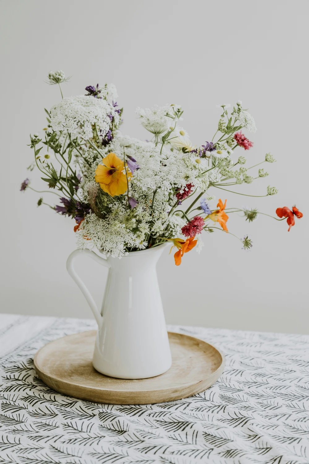 A rustic and charming center display showcases beautiful kitchen table decor ideas for summer with a relaxed, country feel. A white ceramic pitcher, styled like a vintage milk can as a vase, sits in the middle of a round wooden tray. It holds a large, loose bouquet of fresh wildflowers, featuring sprawling white Queen Anne's lace, bright orange and yellow nasturtiums, and small purple blossoms. The arrangement rests on a kitchen table covered with a white tablecloth patterned with dark green botanical fronds, all set against a clean, neutral background that highlights the colorful summer blooms.