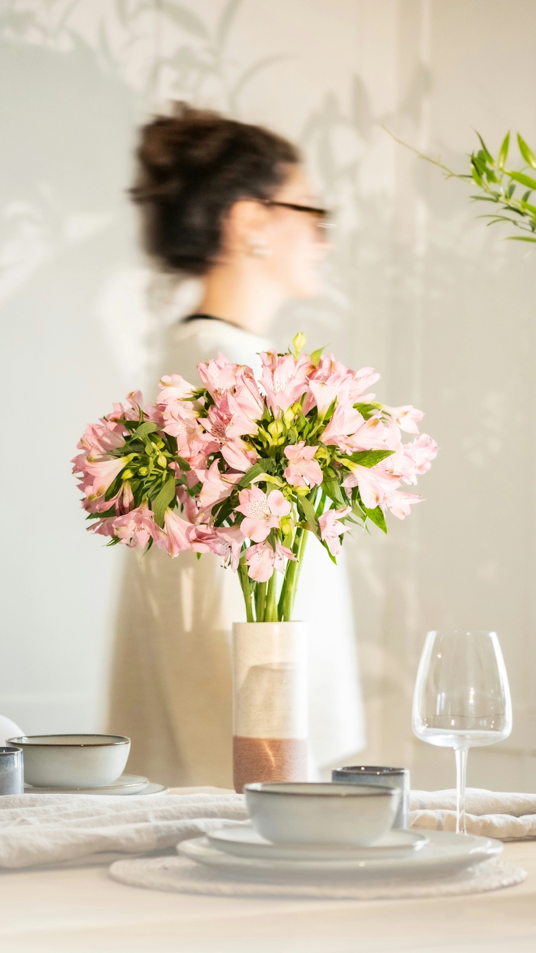 A bright and sunlit dining room highlights delicate kitchen table decor ideas for summer with a fresh, botanical aesthetic. In the center, a tall cylinder ceramic vase with a two-tone white and terracotta base holds a full, vibrant bouquet of pink alstroemeria lilies. The wooden dining table is beautifully laid out with light grey ceramic bowls and plates on round woven placemats, wine glasses, and a textured neutral fabric table runner. In the background, a woman in a white top passes by in a soft blur, while warm sunlight projects artistic leaf shadows onto the clean white wall behind her.
