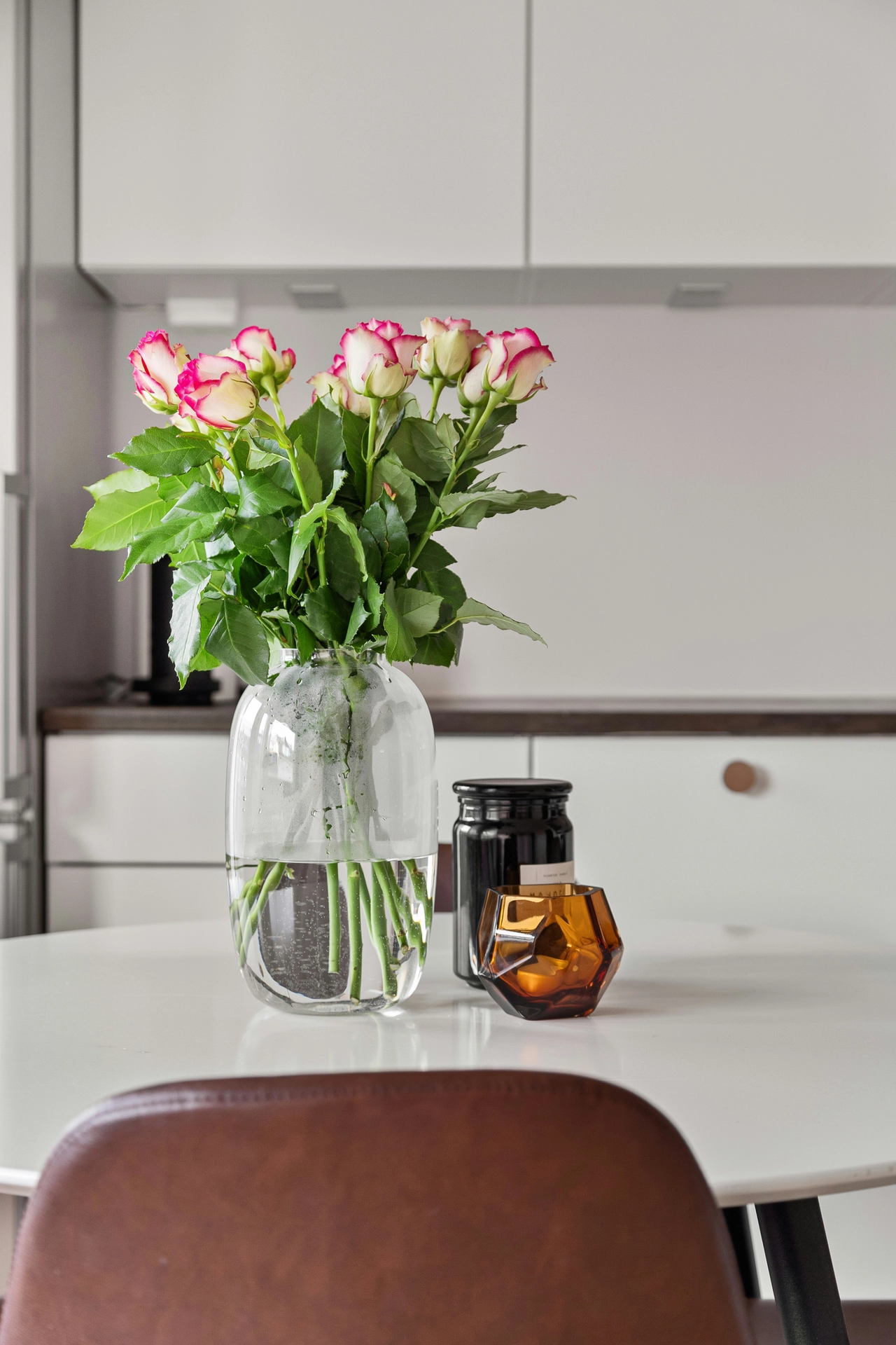 A clean and bright kitchen setting displays modern kitchen table decor ideas for summer with an elegant focal centerpiece. In the middle of a round white dining table sits a beautiful arrangement of roses in a big glass vase, featuring creamy white petals with vibrant magenta pink edges and lush green leaves. Next to the large, clear capsule-shaped vase, a tall black jar and a geometric amber glass candle holder add warmth to the setup. In the background, minimalist white kitchen cabinets with dark countertops complete the crisp, refreshing, and contemporary summer look.