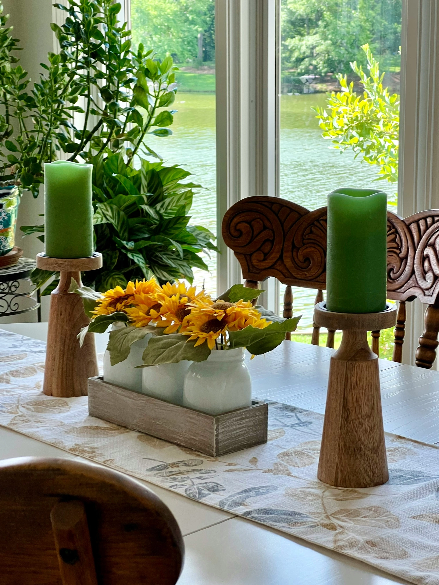 A sunlit dining area showcases cozy, nature-inspired kitchen table decor ideas for summer positioned along a patterned table runner. The centerpiece features a rustic wooden crate holding three small vases framed with candles, each packed with bright yellow artificial sunflowers and green leaves. Flanking this floral arrangement are two tall, wooden pillar candle holders topped with thick green wax candles. In the background, ornate wooden chairs sit before a large window that frames a beautiful, serene view of a sun-drenched lake or river surrounded by lush green trees and indoor potted plants.