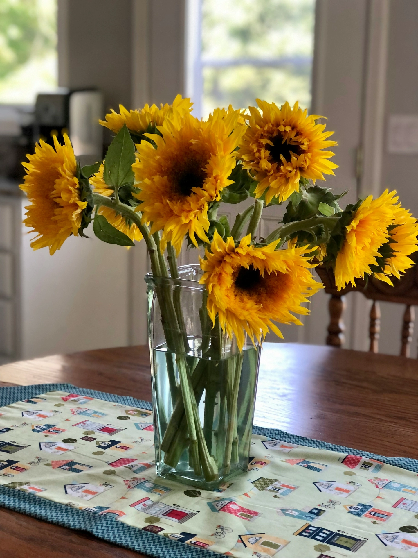 A cheerful and warm home display highlights rustic kitchen table decor ideas for summer atop a dark wooden dining table. Serving as a vibrant centerpiece, a tall, square clear glass vase filled with water holds a large bouquet of bright yellow sun flowers with fuzzy centers and thick green stems. The vase stands on a whimsical fabric table runner patterned with colorful little houses and bordered by a teal chevron trim. In the softly blurred background, a sunny kitchen space with a wooden chair and a bright window creates a cozy, welcoming summer day atmosphere.