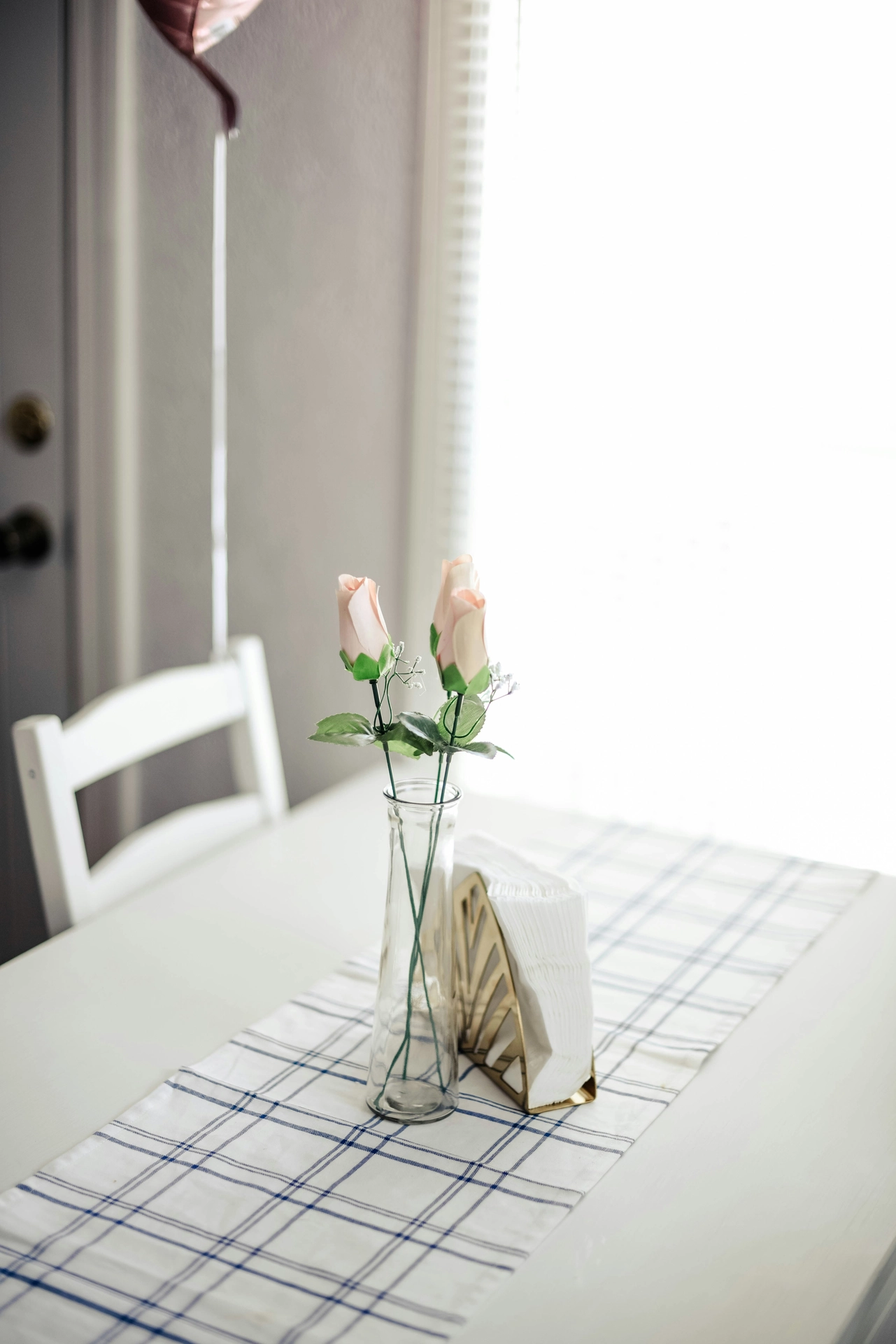A minimalist and bright dining setup showcases clean kitchen table decor ideas for summer with a delicate touch. In the center of a white table, a slender, clear glass bud vase holds two single roses in a soft peach or pale pink hue, accented with tiny sprigs of baby's breath and green leaves. Next to the vase, a modern gold geometric napkin holder is filled with white napkins, all resting on a white fabric table runner with a thin blue plaid or grid pattern. In the background, a white wooden chair and bright, natural light filtering through a window create a calm and airy summer atmosphere.