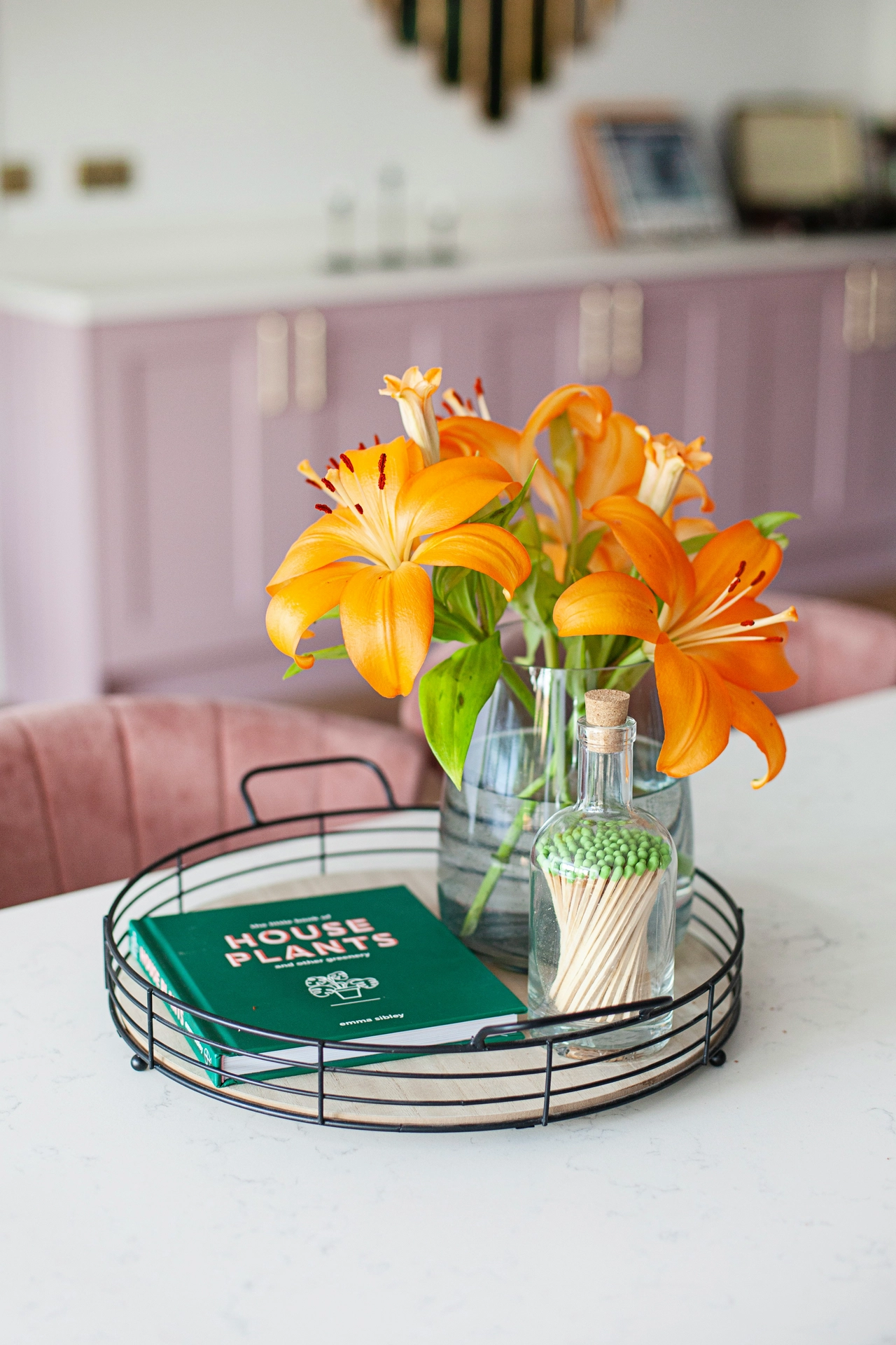A contemporary dining space highlights colorful kitchen table decor ideas for summer arranged neatly on a white marble countertop. The focal centerpiece is styled using a small tray with a black wire rim and a light wood base, which holds a clear glass vase filled with large, vibrant orange lilies. Next to the blossoms, the tray is decorated with a small green book titled 