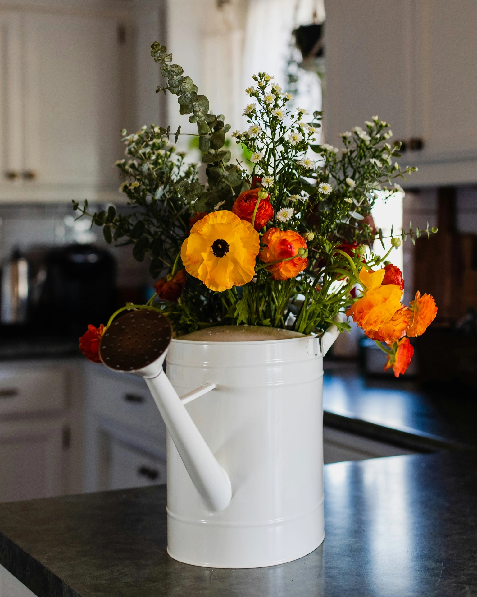 A charming cottage-style display illustrates cozy kitchen table decor ideas for summer set upon a dark countertop. A classic white metal watering can with flowers acts as a unique, rustic vase, overflowing with a bright and colorful seasonal bouquet. The arrangement features large, vibrant orange and yellow ranunculus blooms, tall sprigs of delicate white wildflowers, and textured green eucalyptus stems reaching upward. In the softly blurred background, bright white kitchen cabinets and drawers enhance the clean, sunny, and welcoming farmhouse feel of the scene.