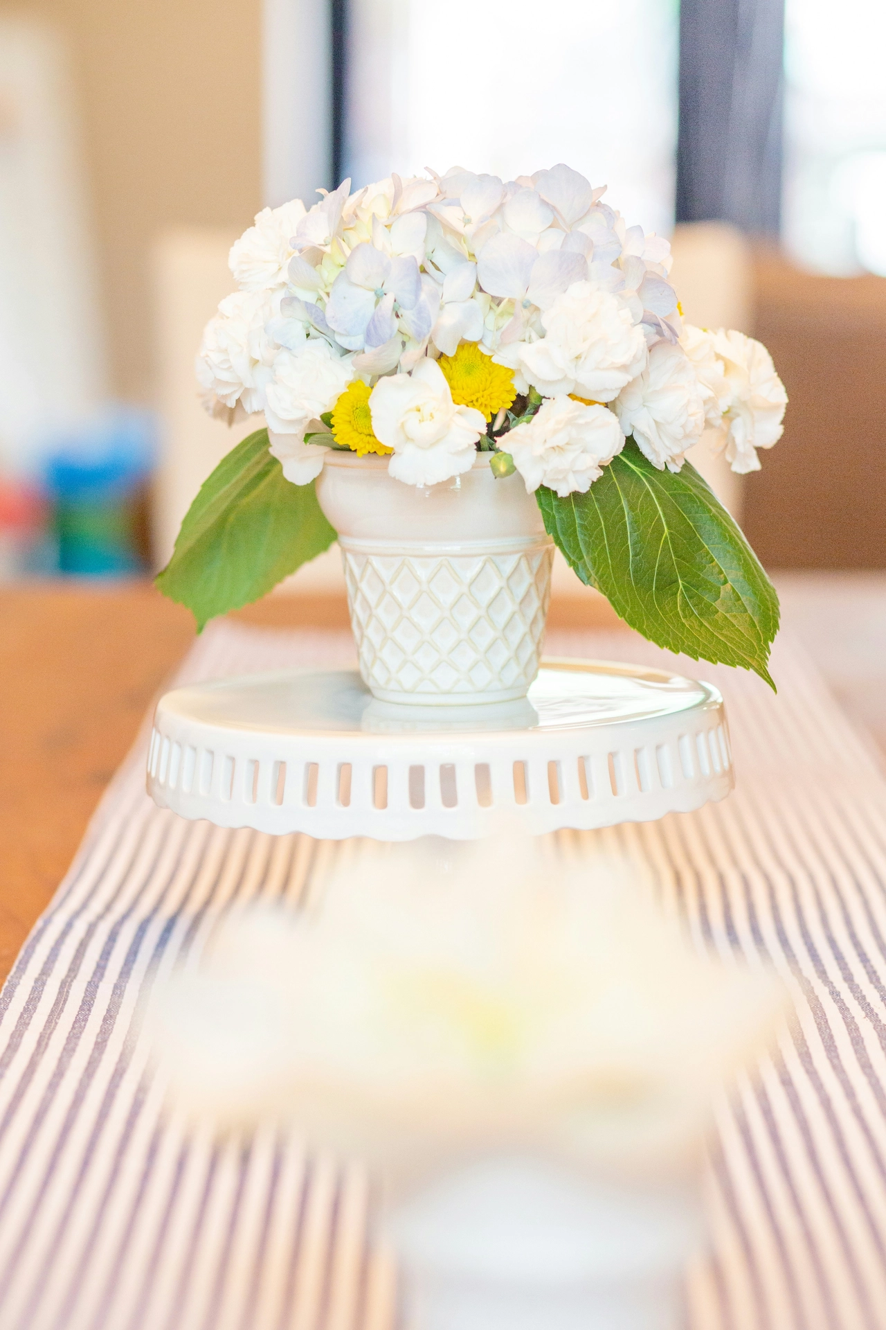 A charming close-up showcases bright kitchen table decor ideas for summer featuring a refreshing coastal color palette. Elevated on a white ceramic pedestal cake stand is a lattice-patterned white cup or small planter acting as a vase. It holds a dense, cheerful bouquet of white and light blue hydrangeas, accented with small yellow chrysanthemums, ruffled white carnations, and broad green leaves. The display stands in the center of a long wooden dining table layered with a classic blue-and-white striped ticking stripe fabric table runner, creating a crisp, inviting, and summery setting.