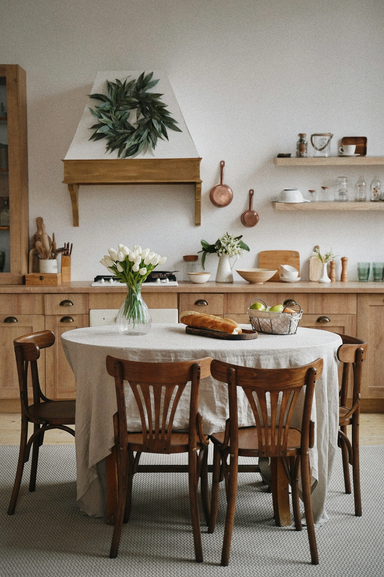 A rustic European-style kitchen beautifully displays charming kitchen table decor ideas for summer on a round dining table covered in a floor-length beige linen tablecloth. The cozy table setup features a ribbed clear glass vase holding fresh white tulips, bread and fruit bowl accents, including a crusty baguette on a wooden board and a wire basket filled with green apples. Four vintage bentwood wooden chairs surround the table on a textured grey rug. In the background, warm light wood cabinetry, open shelving with glass jars, hanging copper pans, and a large green leaf wreath on the white range hood complete the timeless, rustic summer aesthetic.