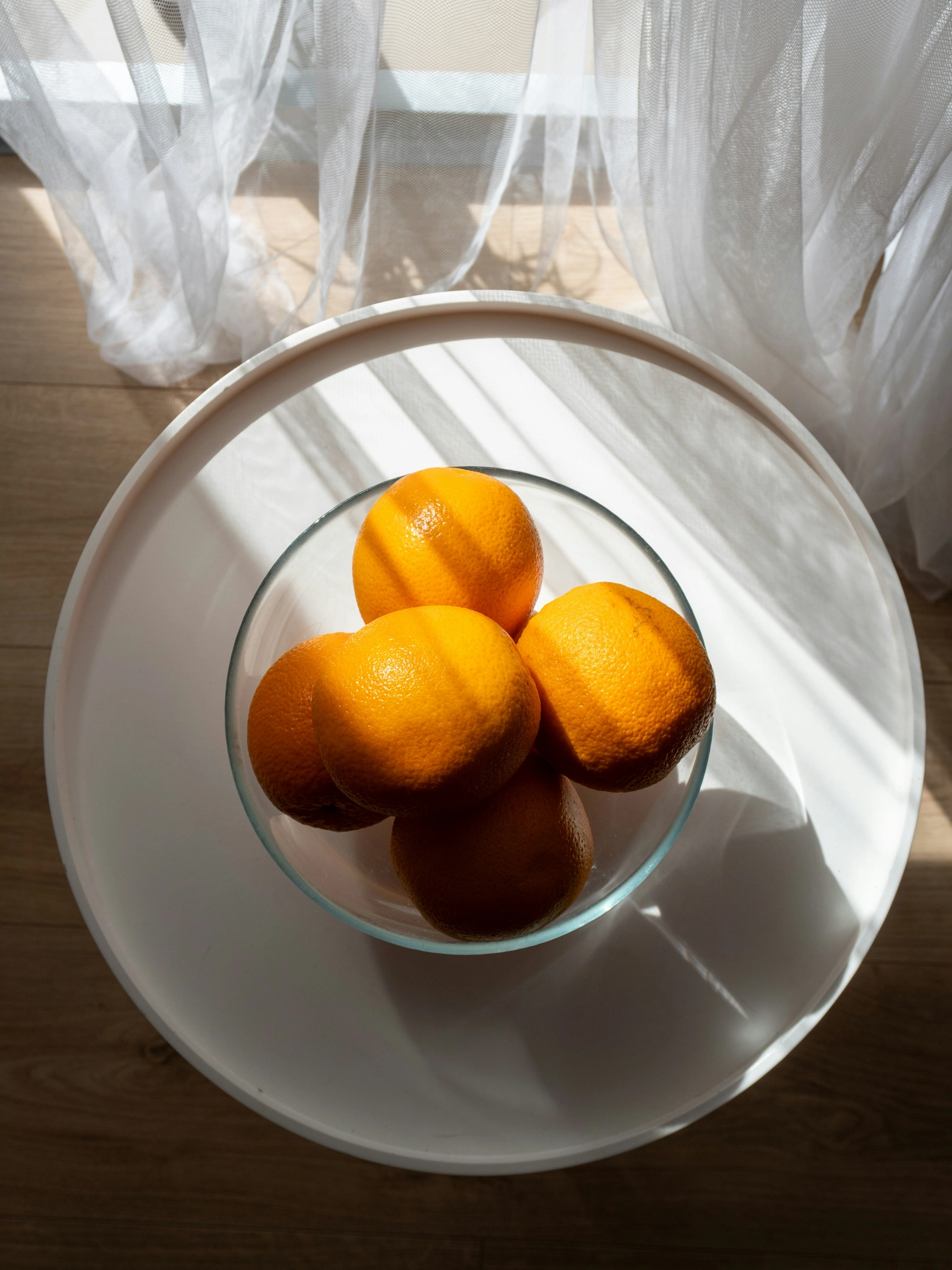 A top-down view captures a bright, sunlit corner showcasing simple yet vibrant summer decor ideas living room styling. A clear glass citrus bowl filled with fresh, bright oranges sits in the center of a round white tray-top side table. Strong summer sunlight streams through delicate, sheer white curtains in the background, casting artistic linear shadows across the table and the rich textured fruit. The combination of the warm orange hues, the clean white surface, and the light wood flooring underneath creates a crisp, refreshing, and distinctly sunny atmosphere.