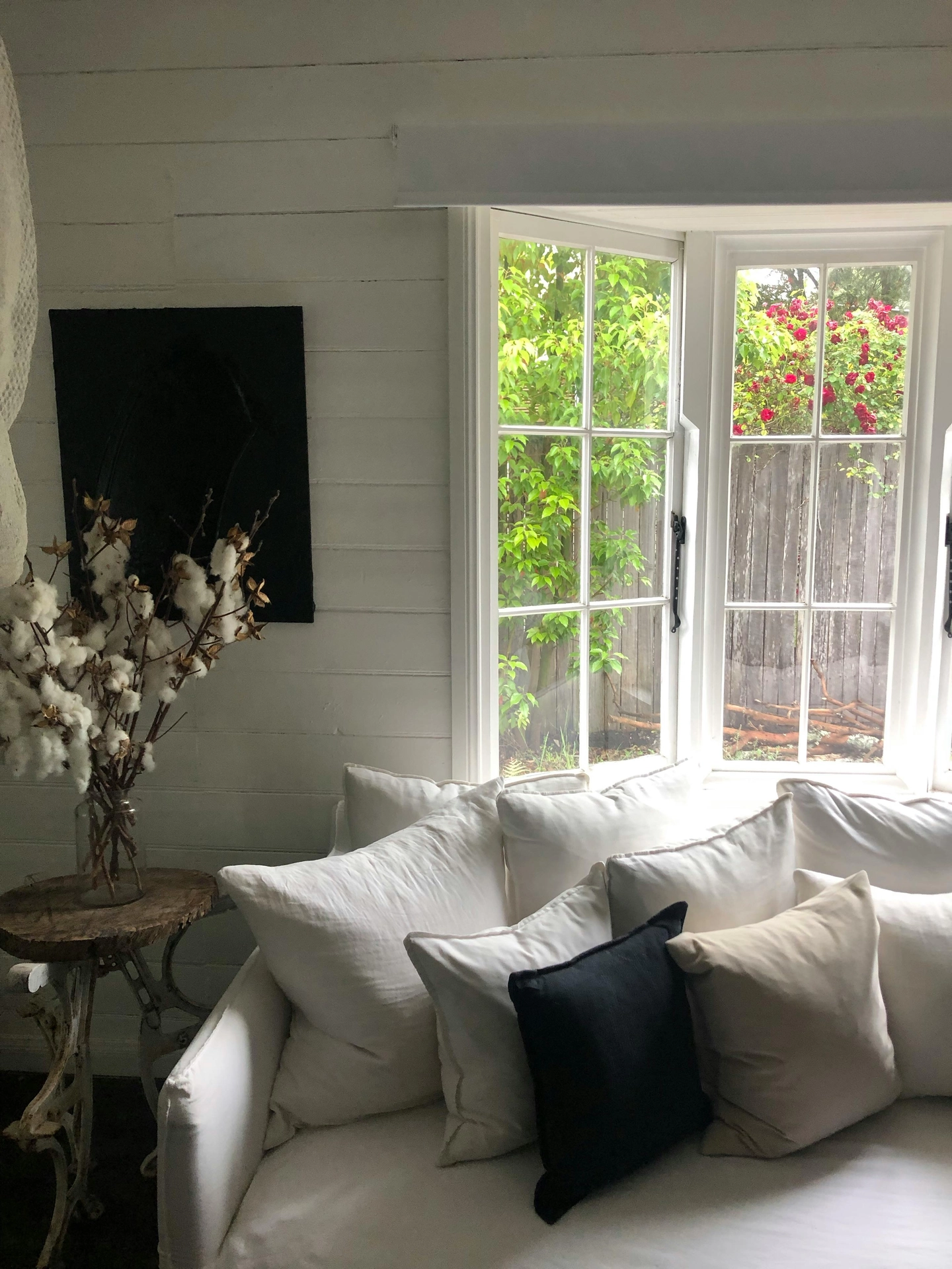 A bright, cozy corner showcases breezy summer decor ideas living room design with a focus on relaxed textures. A deep sofa is fitted with crisp white linen slip covers and layered with numerous large white cushions, accented by a single black and a beige throw pillow. The couch is positioned next to a large white multi-pane bay window that looks out onto a vibrant green garden with blooming red roses. To the left, a rustic wooden side table with an ornate vintage base holds a glass vase filled with fluffy cotton stems, all set against a white horizontal shiplap wall.