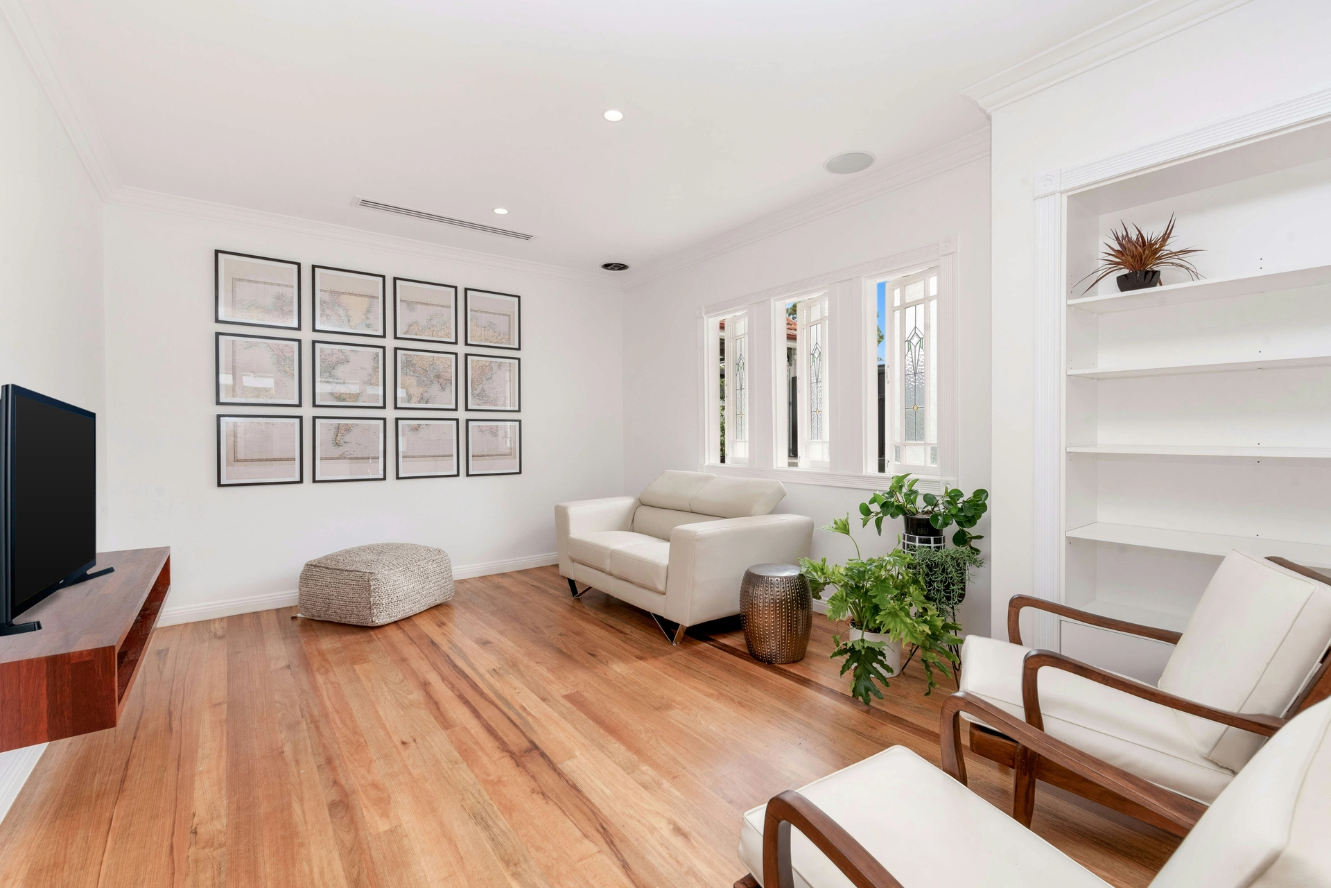 A spacious and airy interior demonstrates minimalist summer decor ideas living room styling by leaving a beautiful hardwood naked floor completely uncovered for a cool, breezy feel. The room is furnished with a sleek off-white leather sofa beneath multi-pane windows and a pair of mid-century wooden armchairs in the foreground. A large gallery wall featuring twelve framed vintage maps serves as a focal point, while a floating wooden television console, a woven pouf, and a cluster of vibrant green potted plants add natural warmth to the crisp white walls and built-in shelving.