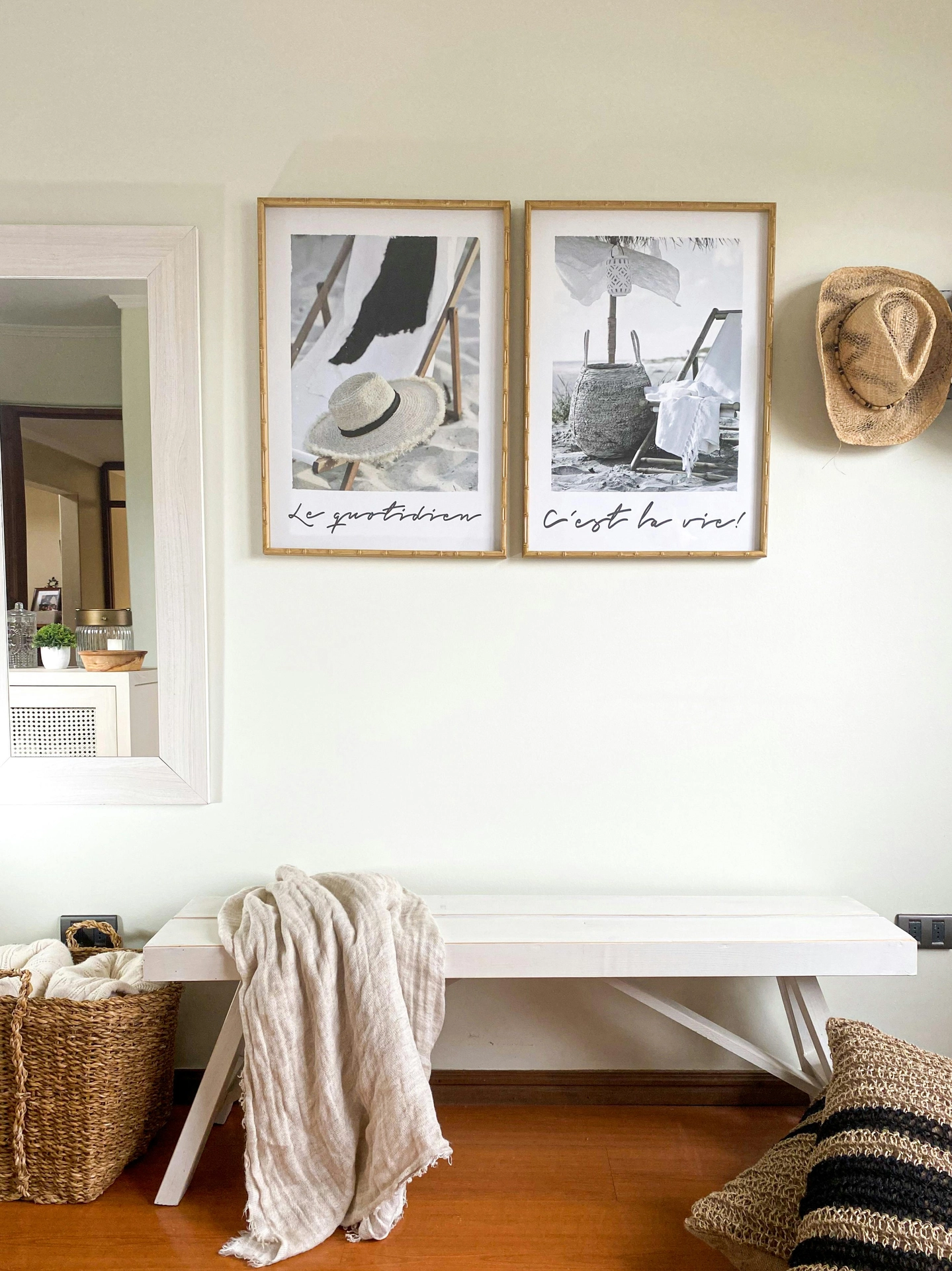 A light cream-colored wall features beachy summer decor ideas living room styling, highlighting a seasonal art swap with two large gold-framed prints. The black-and-white photographs depict a relaxing beach scene, including a straw hat resting on a beach chair and a woven basket under a coastal umbrella, complete with French script cursive underneath. Below the prints, a simple white wooden bench is casually draped with a lightweight, fringed beige linen throw blanket. The coastal, airy look is completed by a woven basket filled with textiles to the left, a large striped jute floor pillow on the hardwood floor to the right, and a straw cowboy hat hanging on the wall.