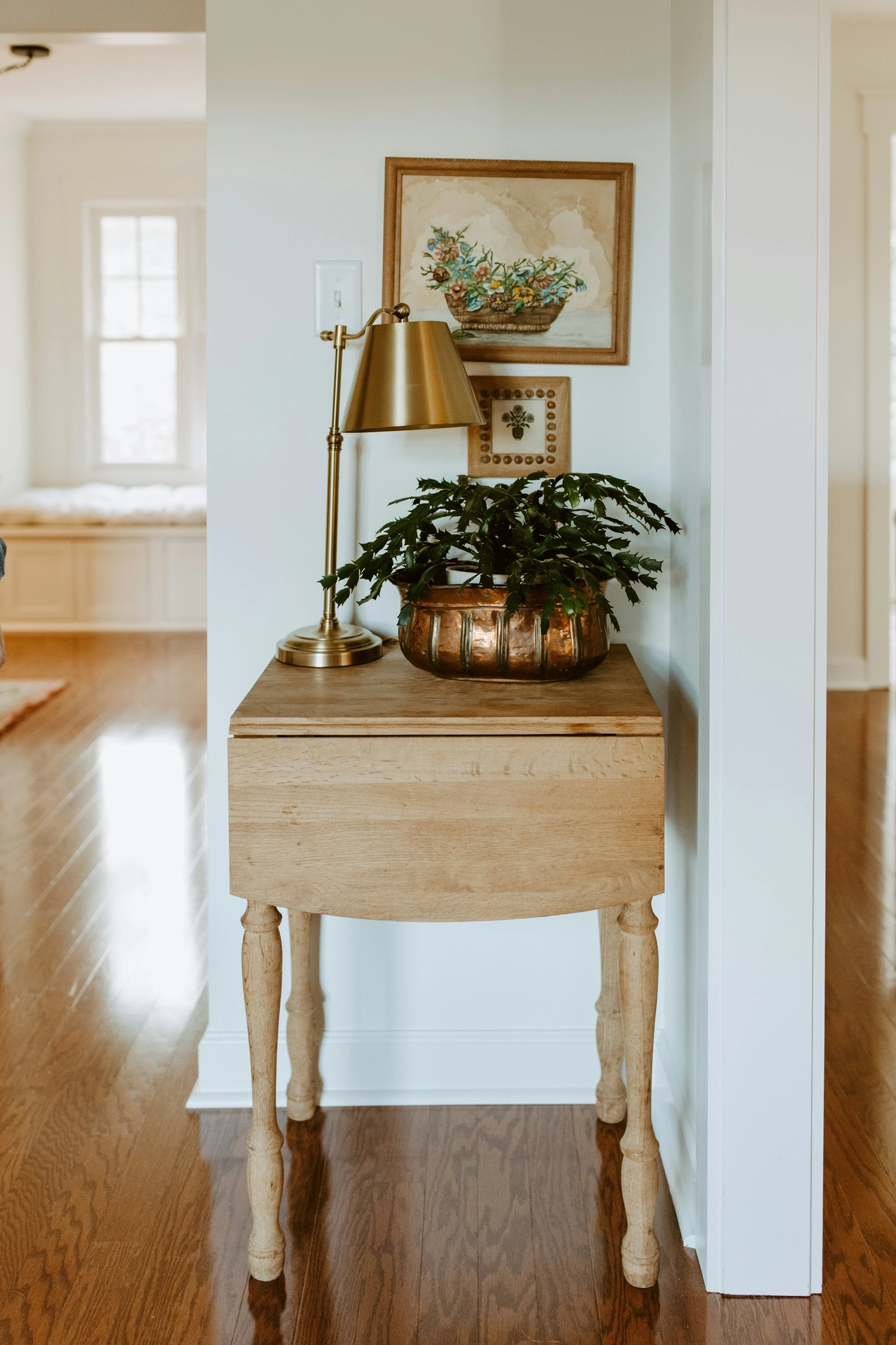 A charming hallway or corner vignette showcases timeless summer decor ideas living room styling with warm metallic touches. A rustic, light-wood drop-leaf side table with turned legs sits on polished hardwood floors against a white wall. The tabletop is styled with elegant brass accents, including a traditional metal desk lamp and a textured copper or brass planter holding a lush, green holiday cactus. Hanging on the wall above the table are two small, vintage-style framed art pieces featuring floral illustrations, adding a classic, cozy touch to the bright and airy home interior.