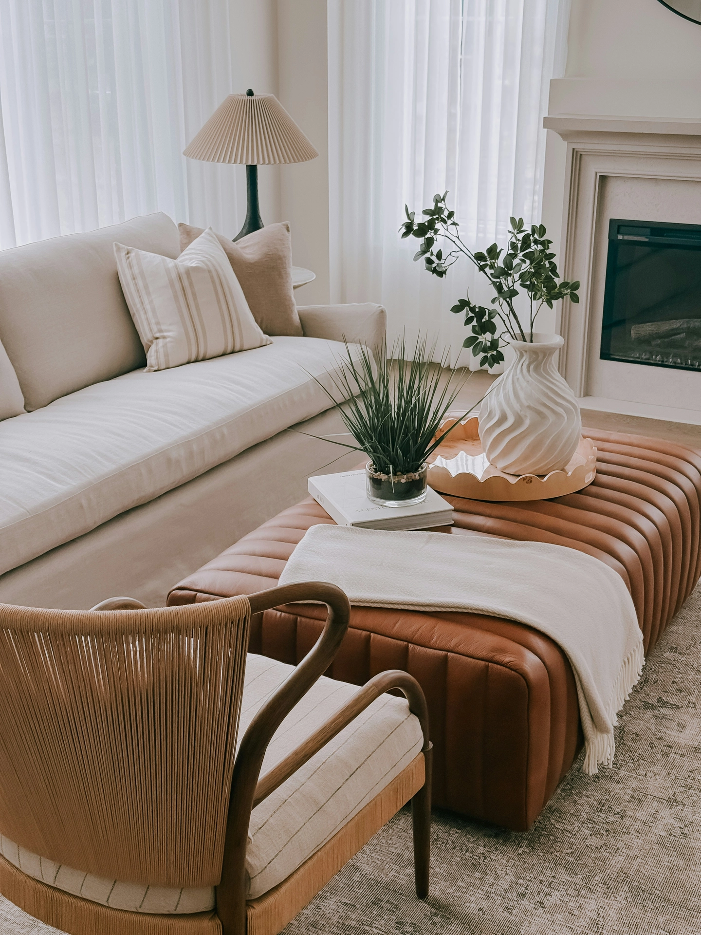 A sophisticated, light-filled space highlights elegant summer decor ideas living room styling with a warm color palette. A large tufted brown leather ottoman serves as a central coffee table, decorated with a scalloped wooden tray that holds a textured white ceramic vase filled with freshly cut green branches. A comfortable beige slipcovered sofa with neutral-toned throw pillows sits next to a woven armchair in the foreground, while a classic fireplace and a pleated lampshade add classic texture. The large windows covered in sheer white curtains filter a soft, bright summer light over the entire contemporary interior arrangement.