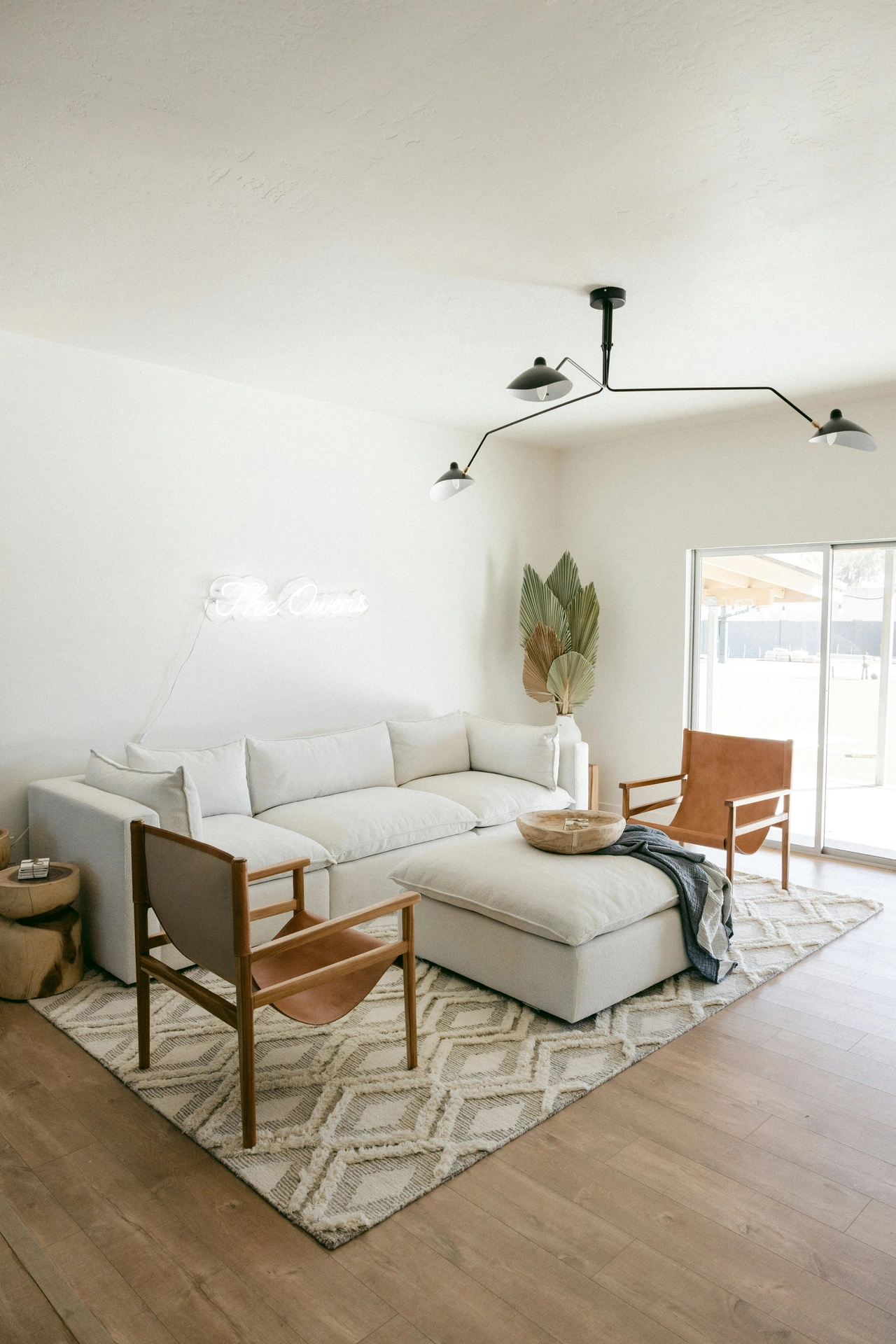 A modern and spacious seating area beautifully executes refreshing summer decor ideas living room styling, emphasizing a palette of clean light colors. A large, off-white sectional sofa and matching oversized ottoman sit on a textured cream diamond-patterned rug over light oak floors. The airy space is furnished with two warm tan leather armchairs with wooden frames, while a large glass sliding door lets in bright afternoon light. A whimsical white neon sign reading 