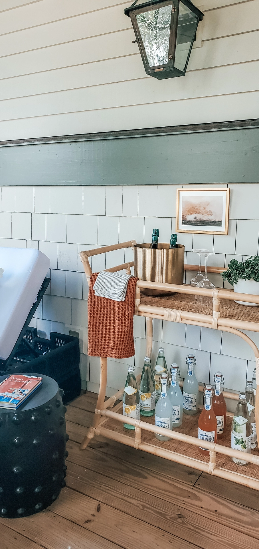 A charmingly styled outdoor porch or sunroom features refreshing summer decor ideas living room styling centered around a two-tiered rattan bar cart. The top shelf of the woven cart is arranged with a polished brass ice bucket holding wine bottles, long-stemmed glassware, a small potted plant, and a rust-orange waffle-knit hand towel draped over the handle. The lower shelf is stocked with a variety of glass soda and lemonade bottles with swing-top caps. Positioned on a natural wooden deck against a shingle-style white wall with green trim, the setup is completed by a black ceramic garden stool, a framed piece of artwork, and a vintage-style hanging wall lantern above.