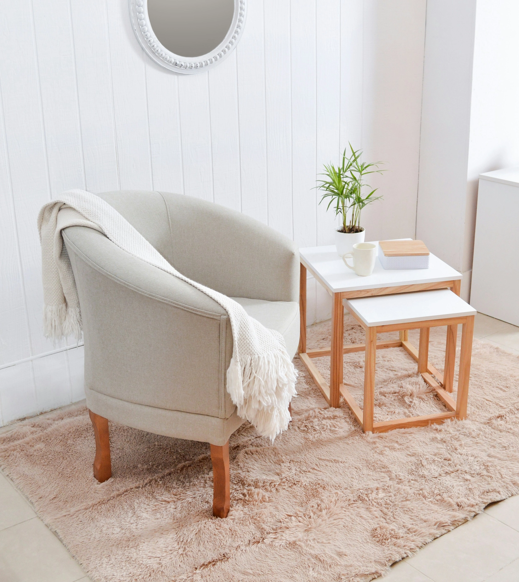 A bright and cozy corner features delightful summer decor ideas living room styling, arranged into a peaceful summer reading nook. A comfortable light beige tub armchair with wooden legs sits on a plush, shaggy tan area rug, adorned with a cream-colored fringed throw blanket draped over its back. Next to the chair, a white nesting table with a light wood frame holds a white coffee mug, a small decorative box, and a potted green plant. The serene setup is placed against a white vertical shiplap wall under a small round mirror with a beaded white frame, creating an inviting spot to relax.