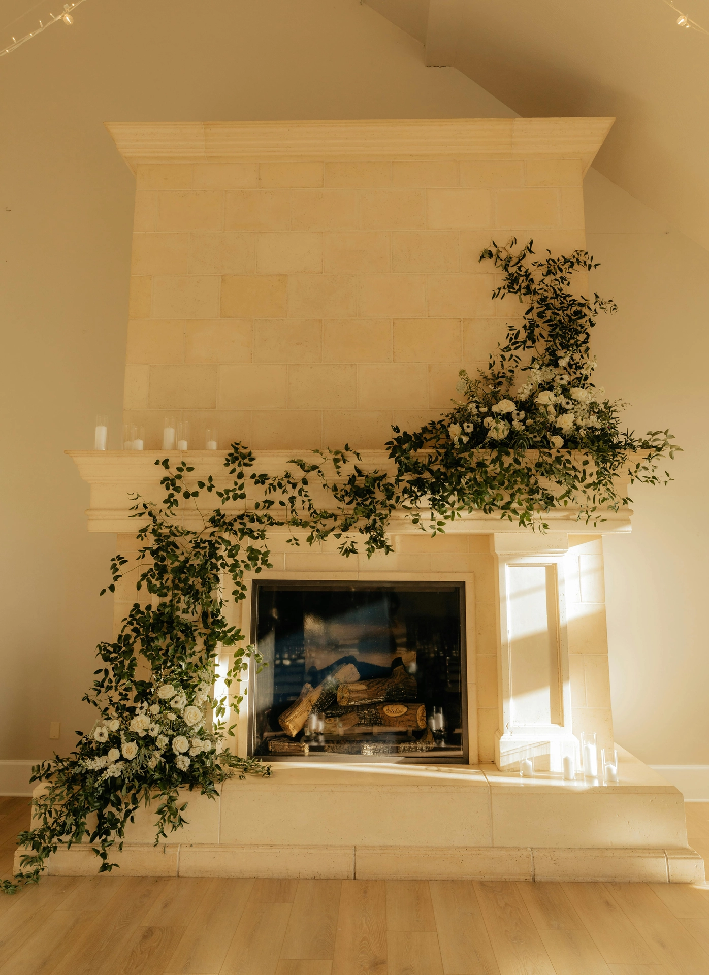 A grand, cream-colored stone fireplace showcases breathtaking floral summer decor ideas living room styling, transforming the hearth into a lush summer fireplace display. Overflowing asymmetric arrangements of fresh green vines and delicate white roses drape elegantly across the mantelpiece and spill down onto the left side of the stone base. The fireplace opening contains neatly stacked logs behind a glass screen, while groups of clear cylinder glass candles are placed on the mantel and hearth. Warm afternoon sunlight streams across the light wood floor, creating a romantic and airy focal point in the room.