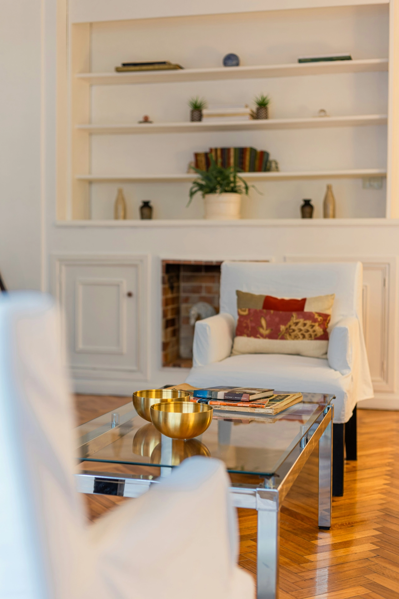 An elegant sitting area features sophisticated summer decor ideas living room styling designed to feel open and airy. In the center, a rectangular chrome coffee table serves as a piece of transparent furniture, with its clear glass top neatly holding two polished brass bowls and a small stack of books. In the background, a white slipcovered armchair accented with a warm-toned patterned pillow sits on polished herringbone wood floors near a classic fireplace hearth. A large built-in white bookshelf is styled with small potted plants, books, and minimal decorative vases, enhancing the clean and breezy look of the room.
