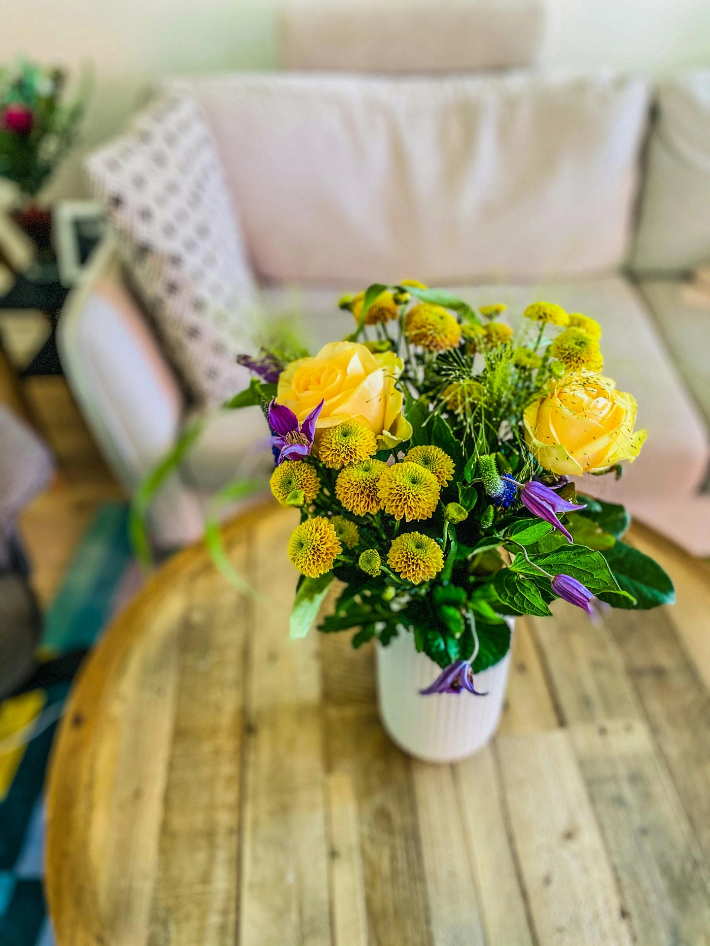 A vibrant bouquet of flowers serves as a beautiful focal point, showcasing simple summer decor ideas living room design. A fluted white ceramic vase sits in the center of an uncluttered coffee table made of natural wooden planks. The cheerful arrangement features large yellow roses, small round golden chrysanthemums, and delicate purple clematis blossoms mixed with fresh green leaves. In the soft-focus background, a light-colored sofa with a patterned throw pillow creates a cozy, inviting, and bright indoor setting that perfectly captures a fresh summer aesthetic.