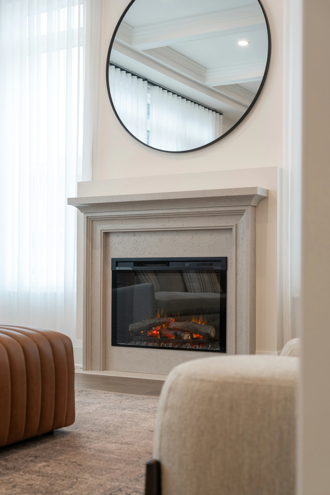 A bright and sophisticated fireplace mantel exemplifies clever summer decor ideas living room styling to expand the look of a space. Mounted directly above a modern light grey stone fireplace is a large, round black-framed mirror, demonstrating how to use mirrors to reflect incoming summer light and create an open, airy feeling. The electric fireplace below features glowing logs behind a glass screen, while sheer white floor-to-ceiling curtains frame the setup. In the foreground, a tufted brown leather ottoman and a textured cream armchair complete the luxurious, comfortable seating arrangement.