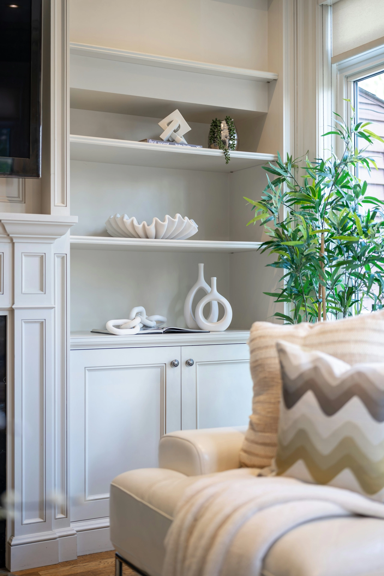 A beautifully curated built-in shelving unit displays elegant summer decor ideas living room styling with a focus on pristine white decor elements. The off-white shelves are artfully arranged with contemporary sculptures, including a ceramic chain link, a pair of circular hollow vases, a large wavy clamshell-inspired bowl, and a geometric interlocking piece. To the right, a lush green potted bamboo plant adds a vibrant touch of nature, while a cream leather armchair with patterned throw pillows sits comfortably in the foreground next to a classic fireplace mantel.