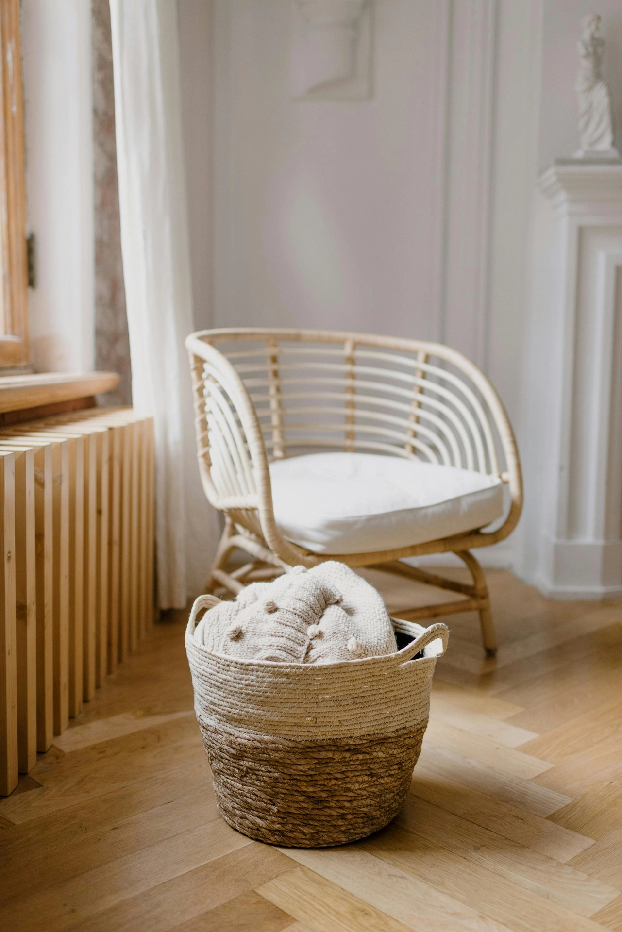 A bright room features elegant summer decor ideas living room styling with an emphasis on natural textures and light tones. In the foreground, one of several large woven baskets with a two-tone design-cream on top and natural jute on the bottom-holds a cozy knitted blanket with pom-pom details. In the soft-focus background, a light-colored bamboo barrel chair with a white seat cushion rests on herringbone hardwood flooring near a sunlit window with a sheer curtain, creating a serene and breezy atmosphere.