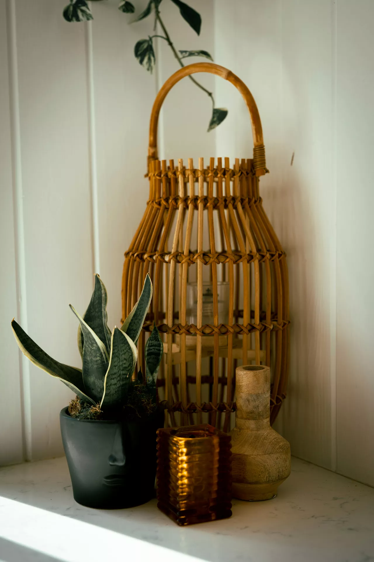 A warm interior corner display showcases inviting lantern summer decor ideas arranged against a clean white shiplap wall. The focal point of the arrangement is a tall, woven light brown bamboo lantern featuring a prominent curved handle and a small glass candle holder visible inside. On the white shelf in front of the lantern sits a dark grey planter shaped like a human face holding a spiky green snake plant, a small square textured amber glass candle holder, and a small turned light wood vase, while a delicate green vine trails down gently from above.