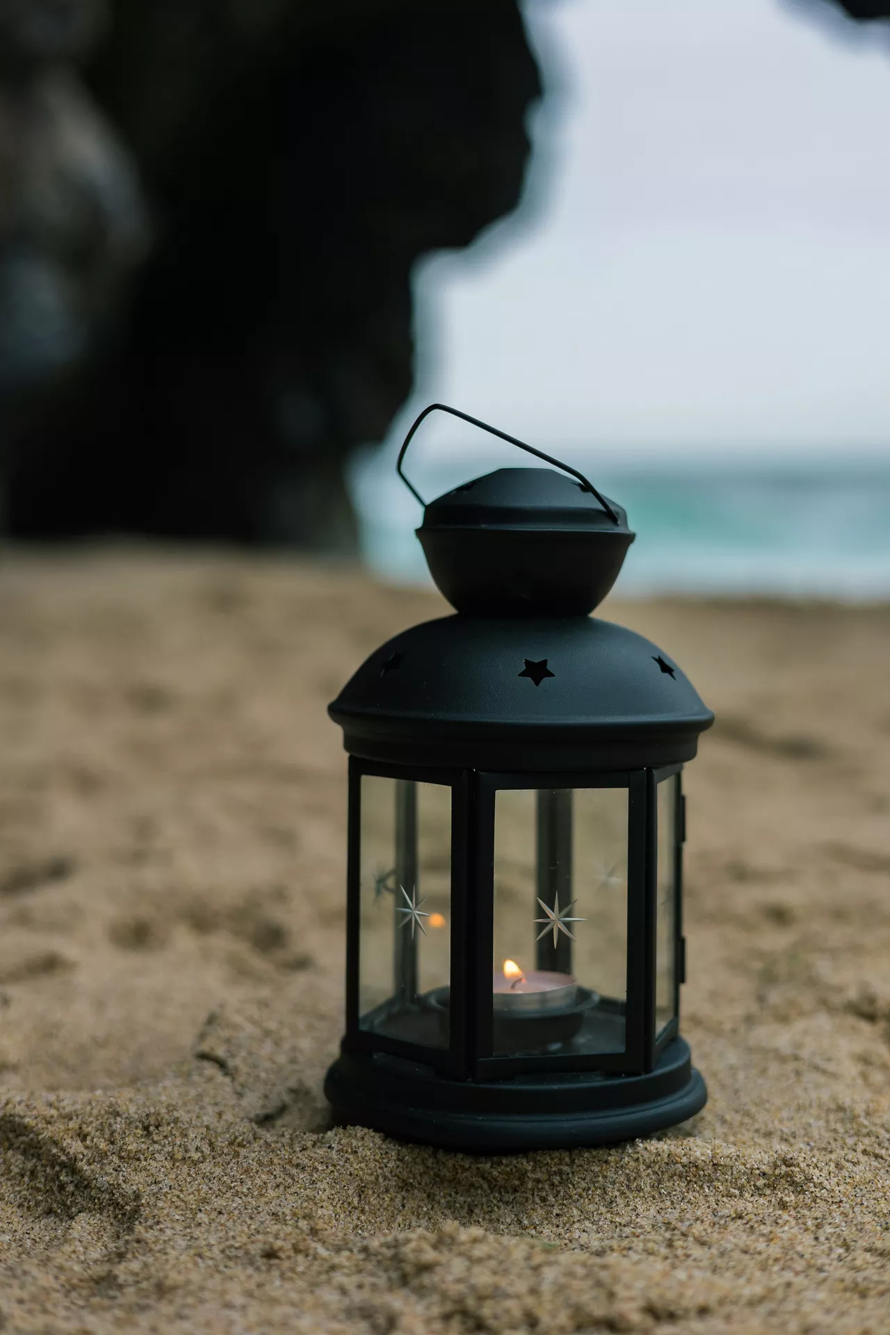 A serene beach setting showcases cozy lantern summer decor ideas with a warm, coastal ambiance. A small black lantern with tea light rests directly on the textured sand, its lit candle throwing a gentle flame inside the glass enclosure. The classic metal lantern is designed with decorative star cutouts on the top dome and a thin wire handle. In the softly blurred background, dark cave-like rock formations and the pale blue ocean waves create a peaceful, overcast seaside atmosphere perfect for a summer evening.