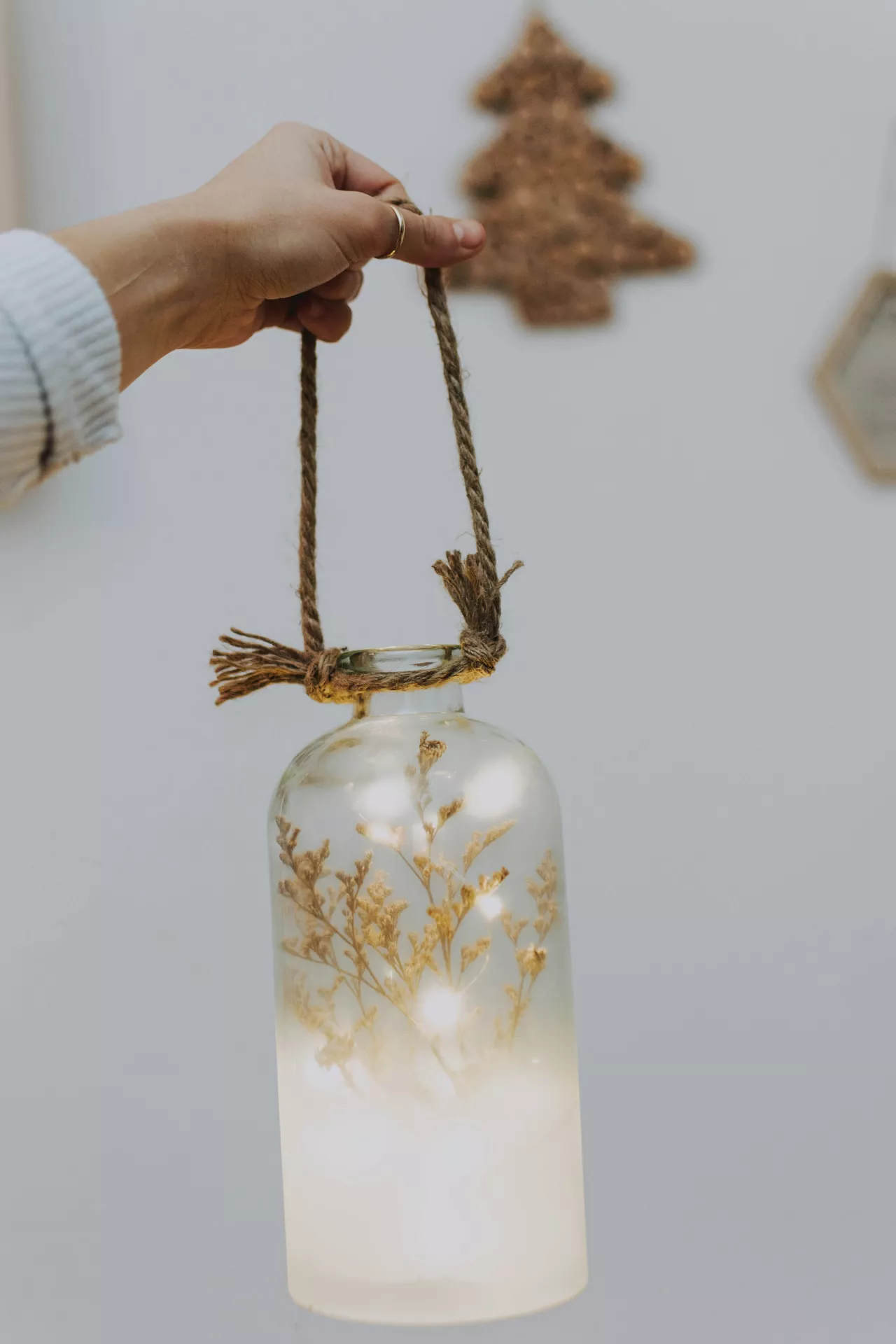 A hand holds up an enchanting example of lantern summer decor ideas against a simple white background. The hand grasps a rustic jute rope handle tied securely around the neck of a clear glass lantern with dried flowers nestled inside. Tiny, warm white fairy lights glow brightly at the base of the bottle, illuminating the delicate, golden dried botanical stems and creating a soft, warm ambiance. In the softly blurred background, a brown textured decoration hangs on the wall, completing the cozy handmade look.