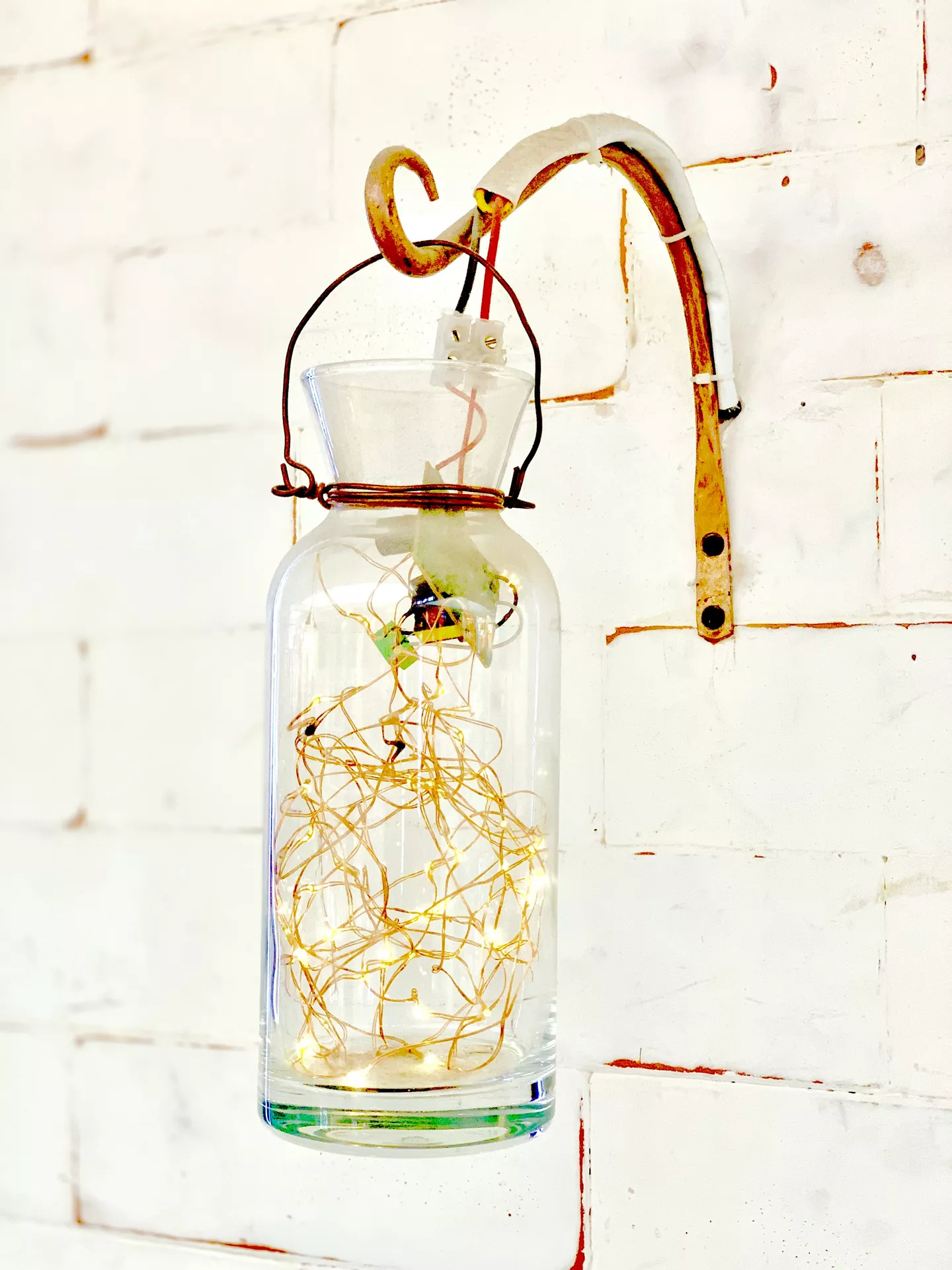 A rustic wall-mounted setup displays industrial lantern summer decor ideas against a white brick wall. A large, clear glass lantern with fairy lights hangs from a curved, weathered metal hook attached to the wall. Inside the glass bottle, a tangled cluster of delicate copper wire string lights glows with warm, tiny points of light near the bottom. The bottle is secured around its neck with wrapped wire and hangs from a thin metal handle, creating a simple yet charming lighting accent for a warm summer evening.