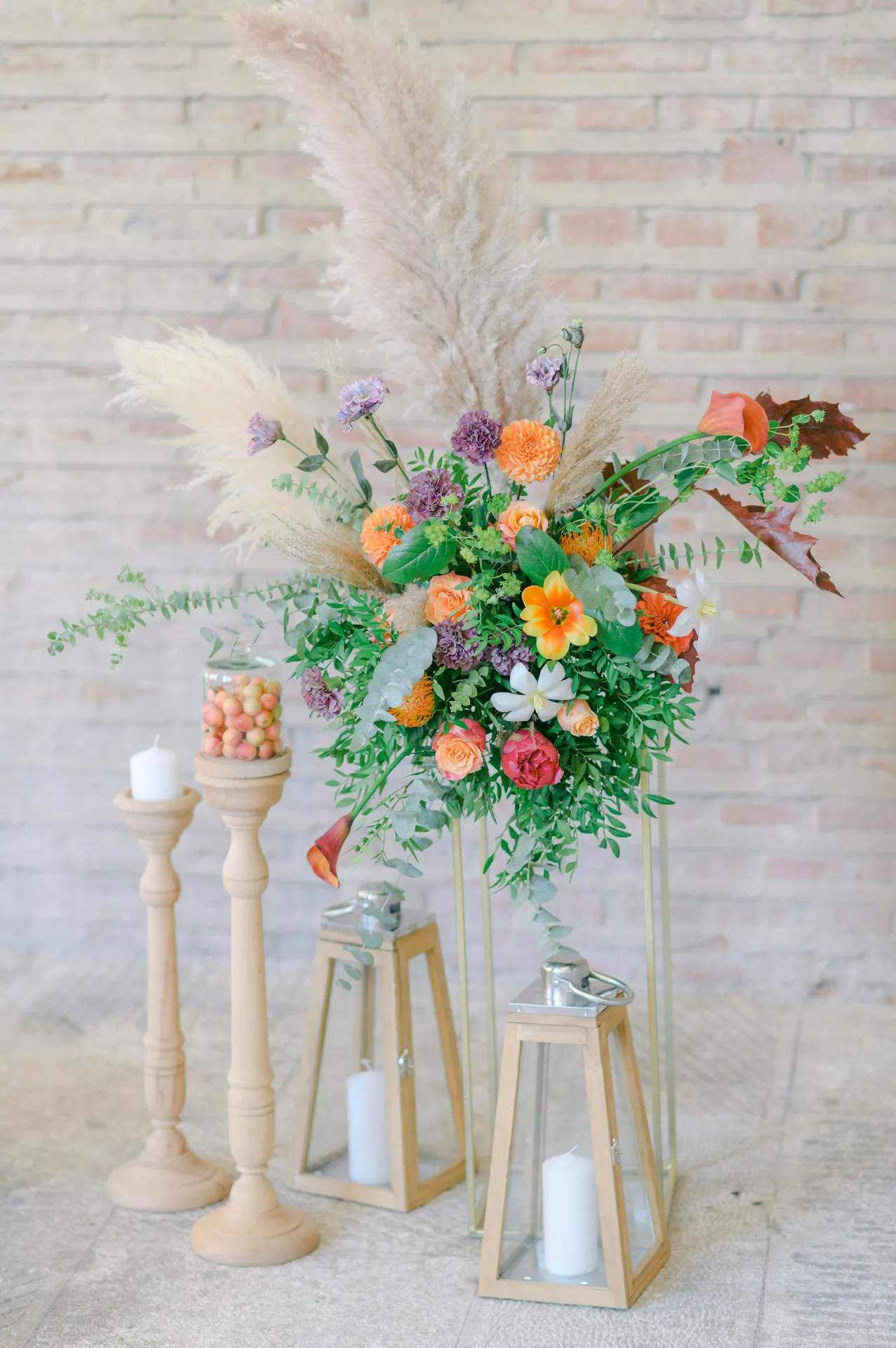 An elegant outdoor display highlights charming lantern summer decor ideas arranged on a rustic dark wooden tabletop. A beautiful lantern group with flowers and candles serves as the centerpiece, featuring three black metal lanterns of varying heights. Inside the lanterns, thick white pillar candles glow with a warm flame, while one lantern is left unlit. The base of the cluster is adorned with an abundant floral arrangement of delicate white laceflowers and fresh green eucalyptus leaves, all set against a softly blurred background of lush summer garden greenery.