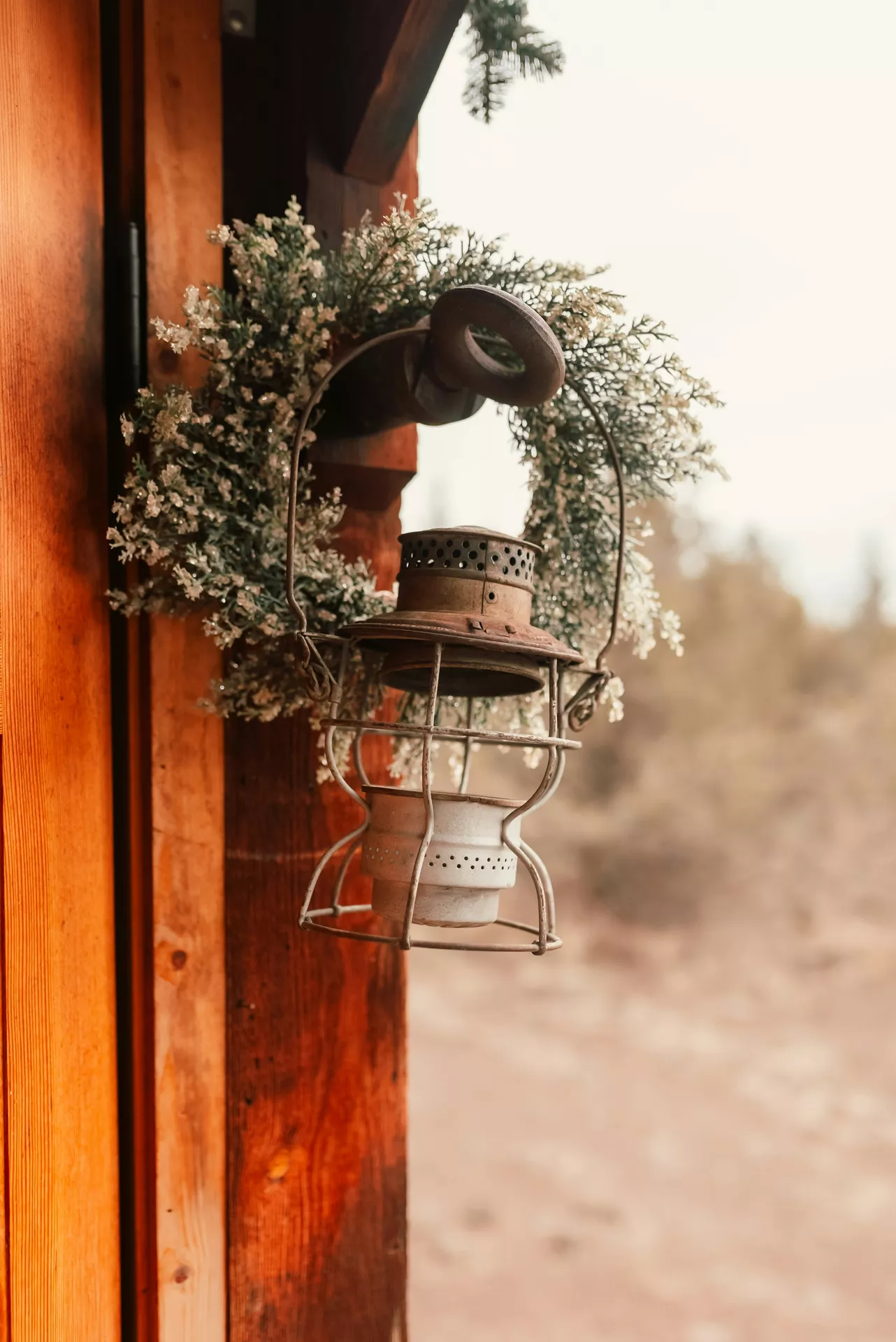 A rustic outdoor scene beautifully presents country-style lantern summer decor ideas hanging from a dark wooden post. An old, unlit vintage metal lantern with flower wreath sits as the centerpiece, featuring a weathered brown top cap and a light grey metal cage base. The rustic lantern hangs from a sturdy metal peg that also supports a lush, delicate circular wreath composed of green frosted pine needles and tiny white wildflowers. In the background, bright natural wood planks of a cabin and a softly blurred, warm-toned natural landscape complete the cozy rustic look.